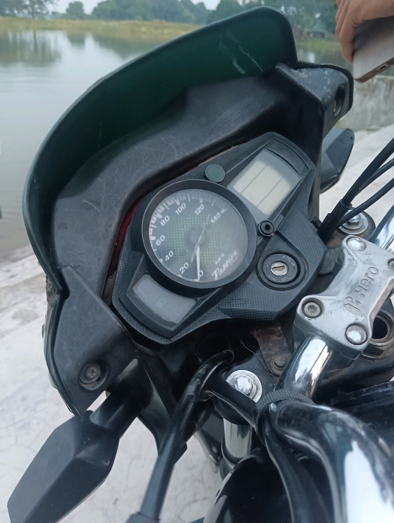 The image captures a close-up view of a motorcycle's dashboard and handlebars. The dashboard features a speedometer with markings up to 140 km/h and a digital display. A key ignition switch is visible below the speedometer, with labels for "ADJUST" and "LOCK". The handlebars are made of chrome, and a component with the "HERO" logo is attached. In the background, a body of water and trees suggest a rural or semi-rural setting, possibly near the city of Tilkar, India. The lighting indicates it is daytime.