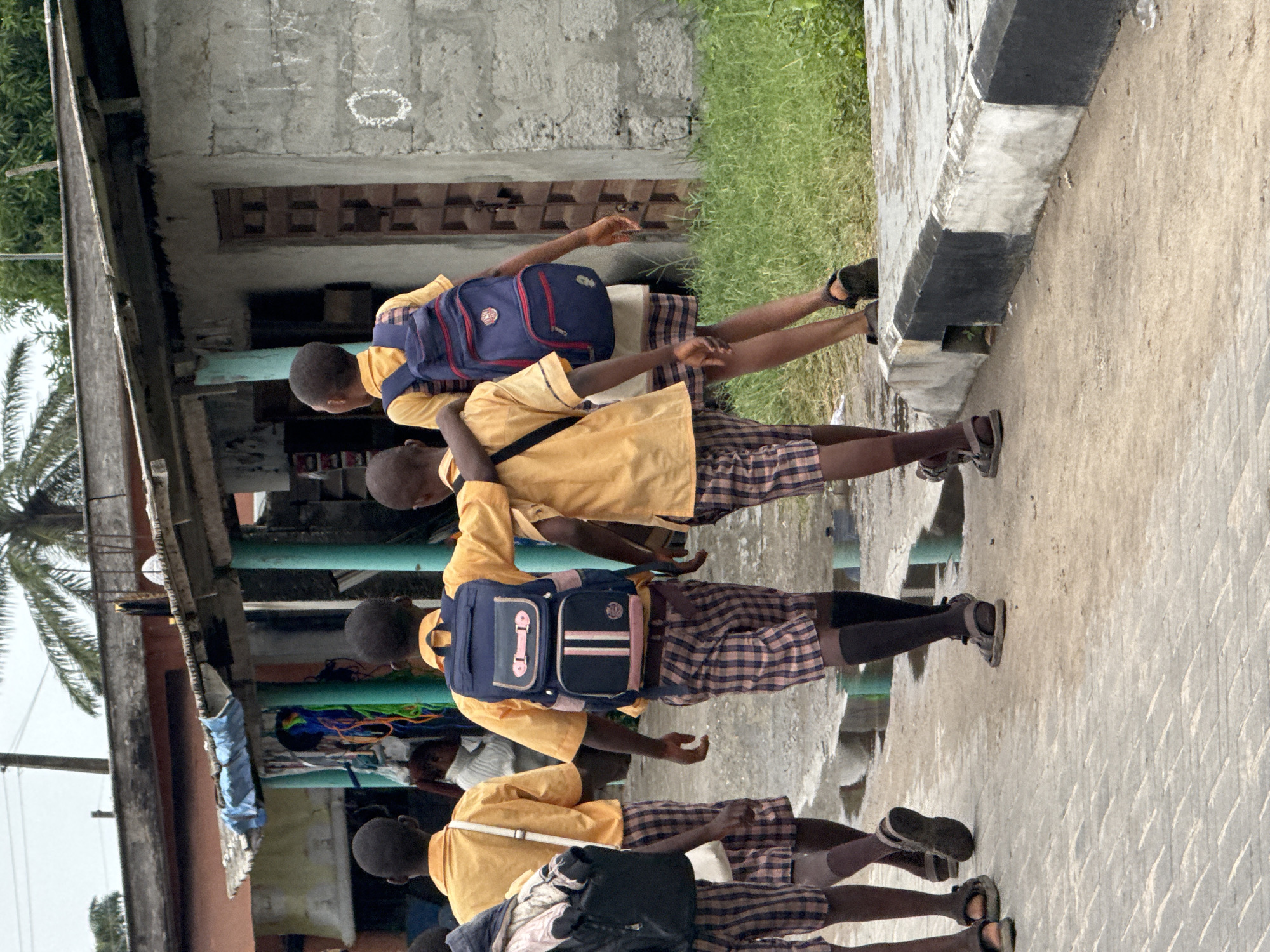 Four children in yellow shirts, plaid shorts, and dark socks, each wearing a backpack, walk along a sidewalk in Badagry, Nigeria. A building with a corrugated roof and green support columns is behind them. A palm tree is visible. The sidewalk has a curb with black and white painted stripes.