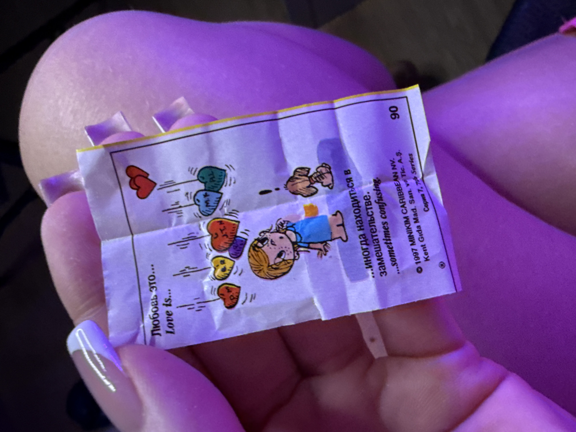 The image shows a hand holding a small, crinkled paper. The paper has a cartoon illustration of a girl standing with heart-shaped balloons floating above her. The text on the paper is in both Russian and English, reading "Любовь это... Love is... ...иногда находиться в замешательстве. ...sometimes confusing." and also includes the copyright information "© 1997 MINIKIM CARIBBEAN NV. Kent Gıda Mad. San. ve Tic. A.Ş. Серия 7, 76 Series" The number "90" is printed in the top right corner of the paper. The hand holding the paper has a light complexion and a white and lilac French manicure. The background features skin and an indeterminate dark area. The overall lighting is purple.
There is nothing about the location in the city of Batumi, Georgia.