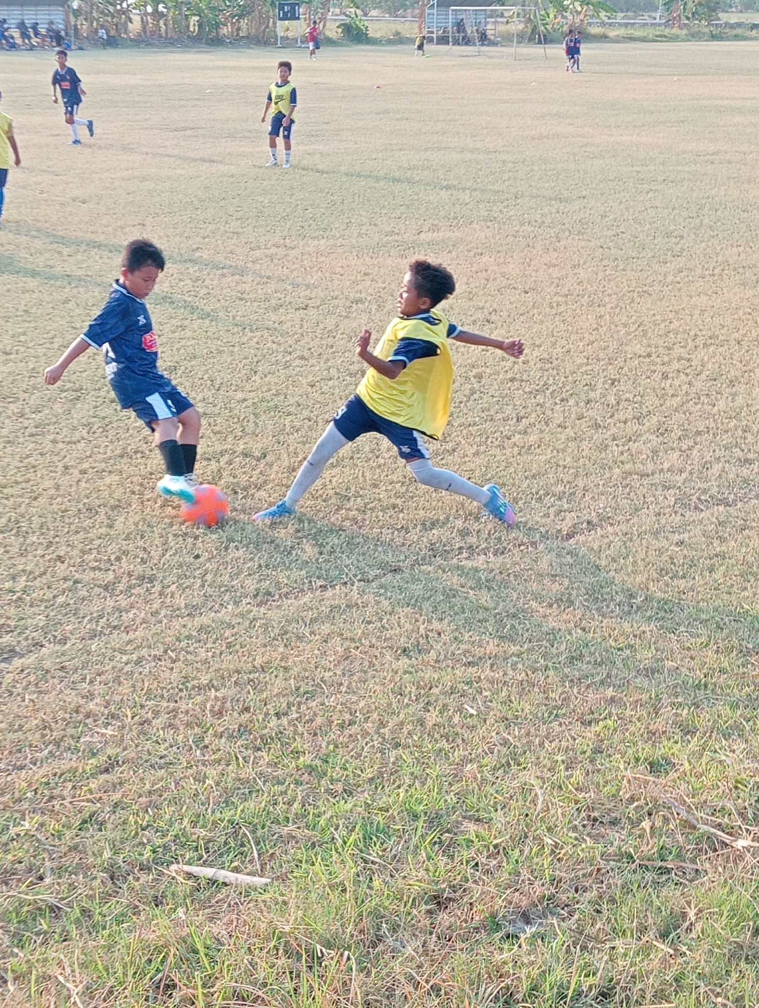 The image depicts a soccer game being played on a dry, grassy field. Two young boys are the primary focus. One boy, wearing a dark blue jersey with white accents and dark shorts, is kicking an orange soccer ball. His left leg is extended, and his right leg is bent as he makes contact with the ball. The other boy, wearing a yellow vest over a blue jersey and dark shorts, is in motion, appearing to defend or attempt to intercept the ball. He has his arms outstretched, and his legs are in a dynamic pose. The background shows more children in soccer attire, some standing and some in motion, on the field. A goalpost is visible in the distance, along with trees and what appears to be a building or structure. The lighting suggests it is daytime, possibly late afternoon due to the warm, low-angled sunlight. The overall environment is an outdoor sports field. The location is identified as Kenep, Indonesia.