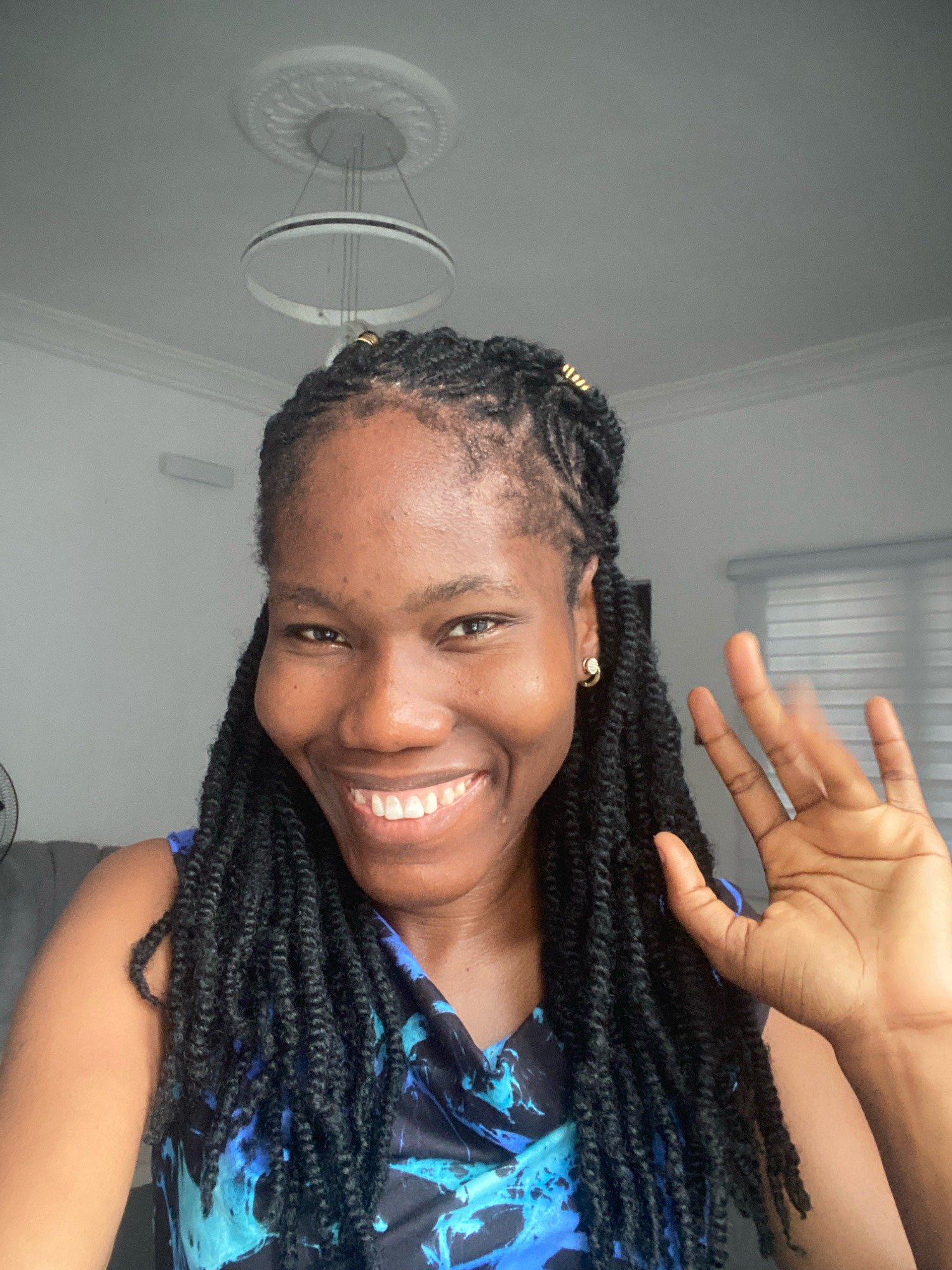 A medium shot photograph captures a Black woman with braided hair, smiling and waving at the camera. She is positioned in the foreground, with her face taking up a significant portion of the frame. Her expression is joyful, with her eyes crinkled and teeth showing in a wide smile. Her right hand is raised, with fingers slightly spread, in a clear waving gesture. She is wearing a dark blue top with a vibrant blue and black abstract pattern. A subtle gold hoop earring is visible in her left ear.
The background suggests an indoor setting, likely a home. A modern circular pendant light fixture hangs from the ceiling, adorned with decorative molding. The walls are painted a neutral color, and a window with blinds is visible to the right, indicating natural light entering the room. To the left, a portion of a dark sofa and what appears to be a fan can be glimpsed, hinting at a living space. The overall impression is one of a casual and welcoming environment. The presence of modern interior design elements, such as the ceiling light and molding, suggest a contemporary home. The lighting is soft and even, contributing to the overall warm and friendly tone of the photograph. The image appears to have been taken in the city of Ado, Nigeria, based on the context provided.