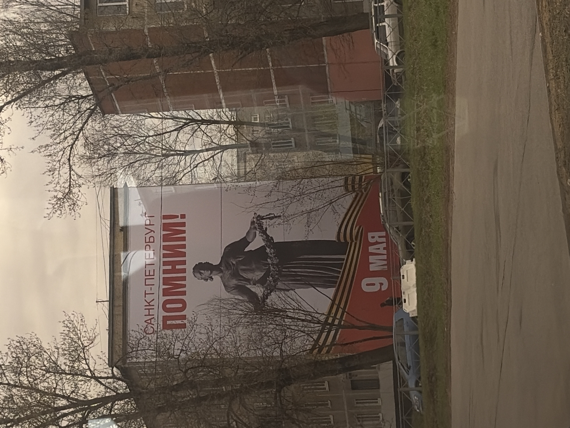 The image shows a billboard in Saint Petersburg, Russia, commemorating Victory Day on May 9th. The billboard features a statue holding a wreath, and is decorated with the Ribbon of Saint George. The text on the billboard reads "Saint Petersburg, We Remember!" in Russian. The background includes apartment buildings and trees without leaves, suggesting early spring. A road runs along the side.