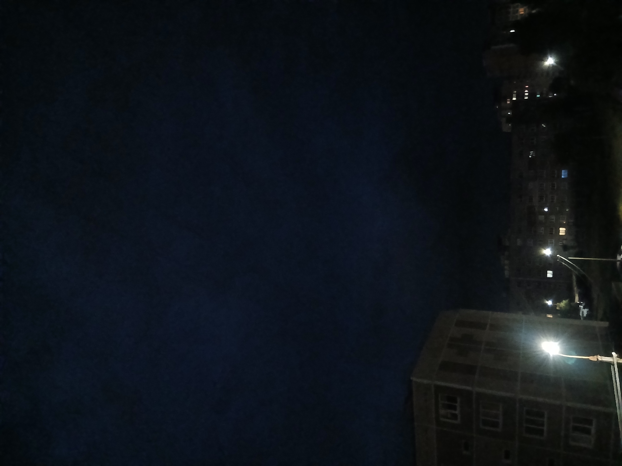 The image depicts a nighttime scene in Chaikovsky, Russia, characterized by a deep blue, cloud-filled sky. In the foreground on the left, a tall, rectangular, light-colored building is visible, with multiple floors and windows. A bright streetlamp illuminates the area in front of this building, casting a strong glow. To the right of this building, a darker, longer apartment building stretches across the middle ground. Numerous windows of this building are lit from within, suggesting occupancy. Silhouetted trees partially obscure the view of the apartment building. In the distance, more buildings are faintly visible, also with illuminated windows. The overall impression is of a residential urban environment at night.
