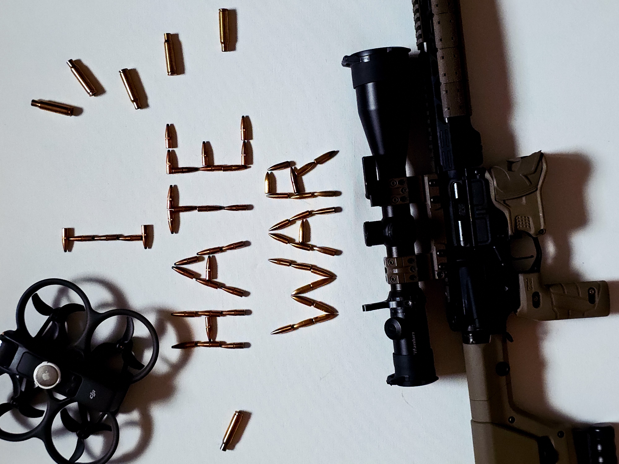 The image displays a still life composition featuring a rifle, a drone, and ammunition arranged on a plain, light-colored surface, likely indoors. A long rifle, appearing to be an AR-style firearm, is positioned on the right side of the frame, angled diagonally. It has a black receiver and barrel, a large black scope (with "Titanium" visible on it) mounted on top, and tan/FDE colored furniture including the stock, pistol grip, and handguard.
To the left of the rifle, numerous bullet casings and pointed projectiles are meticulously arranged to spell out the phrase "I HATE WAR". The metallic copper and brass tones of the ammunition contrast sharply with the white background.
In the lower-left corner, a black DJI Avata drone is visible, showcasing its protective propeller guards and the central camera gimbal. The "DJI" logo is clearly discernible on the drone's body.
The scene is illuminated from one side, casting prominent shadows from all objects, which adds depth and a dramatic effect to the composition. The deliberate arrangement of these objects, particularly the juxtaposition of firearms and a drone with the clear message "I HATE WAR", creates a powerful and thought-provoking statement on conflict.