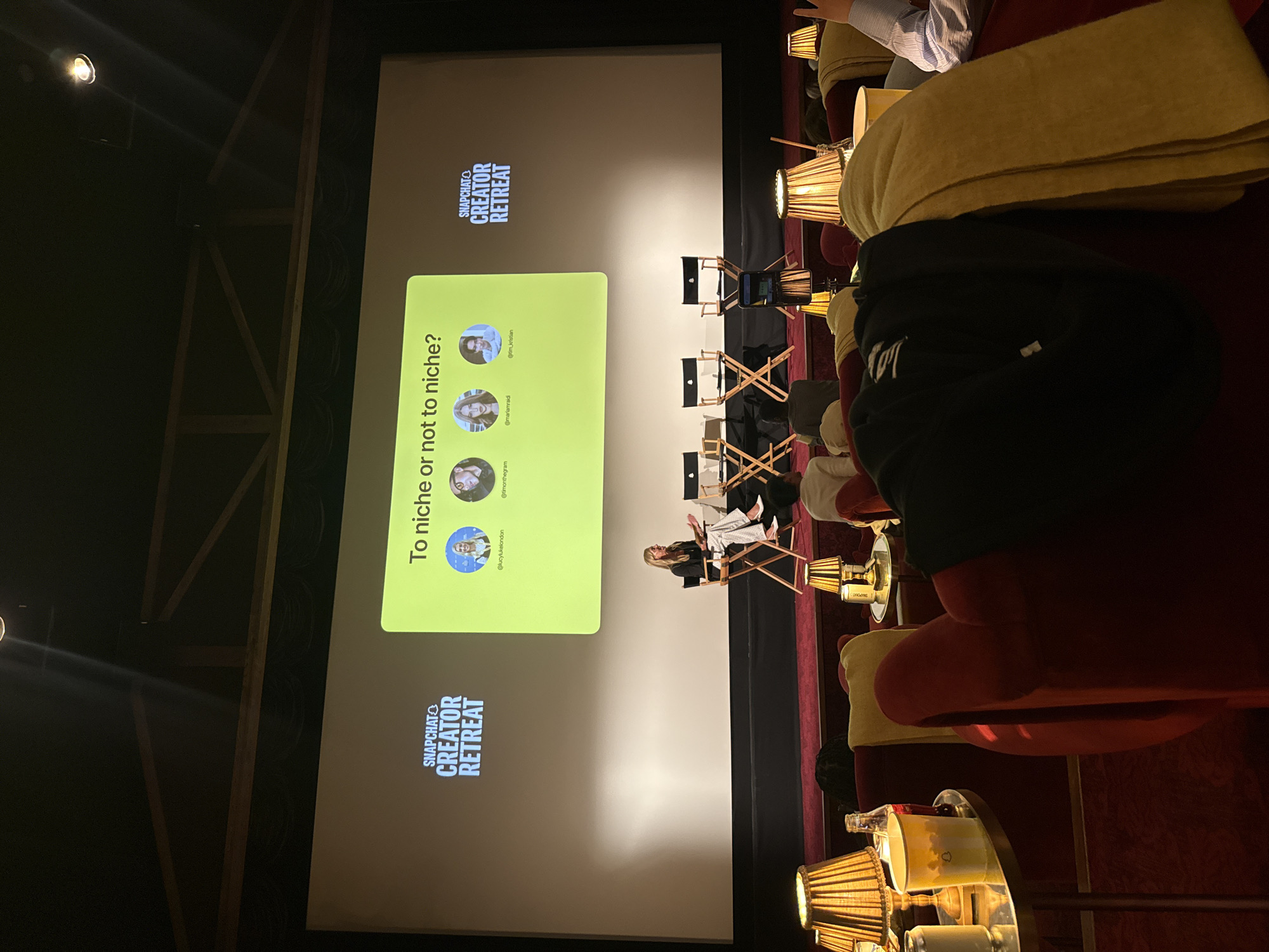 An indoor event, likely a conference or panel discussion, is captured from an audience perspective. A large screen dominates the front, displaying content for a "SNAPCHAT CREATOR RETREAT." The screen features the question "To niche or not to niche?" in a light green box, along with four profile pictures and associated social media handles: "@lucylukelondon", "@timonthegram", "@mariamraidi", and "@tim_kristian".
On a platform in front of the screen, a person is seated in a director's-style chair, facing the audience. Several other empty director's chairs are arranged, suggesting a panel is either ongoing or about to begin. The room has a theater-like ambiance with dim lighting, and spotlights illuminate the stage area.
Audience members are visible in the foreground and mid-ground, seated in comfortable red upholstered chairs. Some chairs have side tables with decorative, possibly wicker or rattan, lamps that emit a warm glow. A yellow blanket is draped over one of the chairs, and a bottle and mug are visible on a side table. One audience member is seen from behind, wearing a dark hoodie, while another wears a striped shirt. The overall setting suggests a focused, intimate event. The location context indicates West Oxfordshire, United Kingdom.