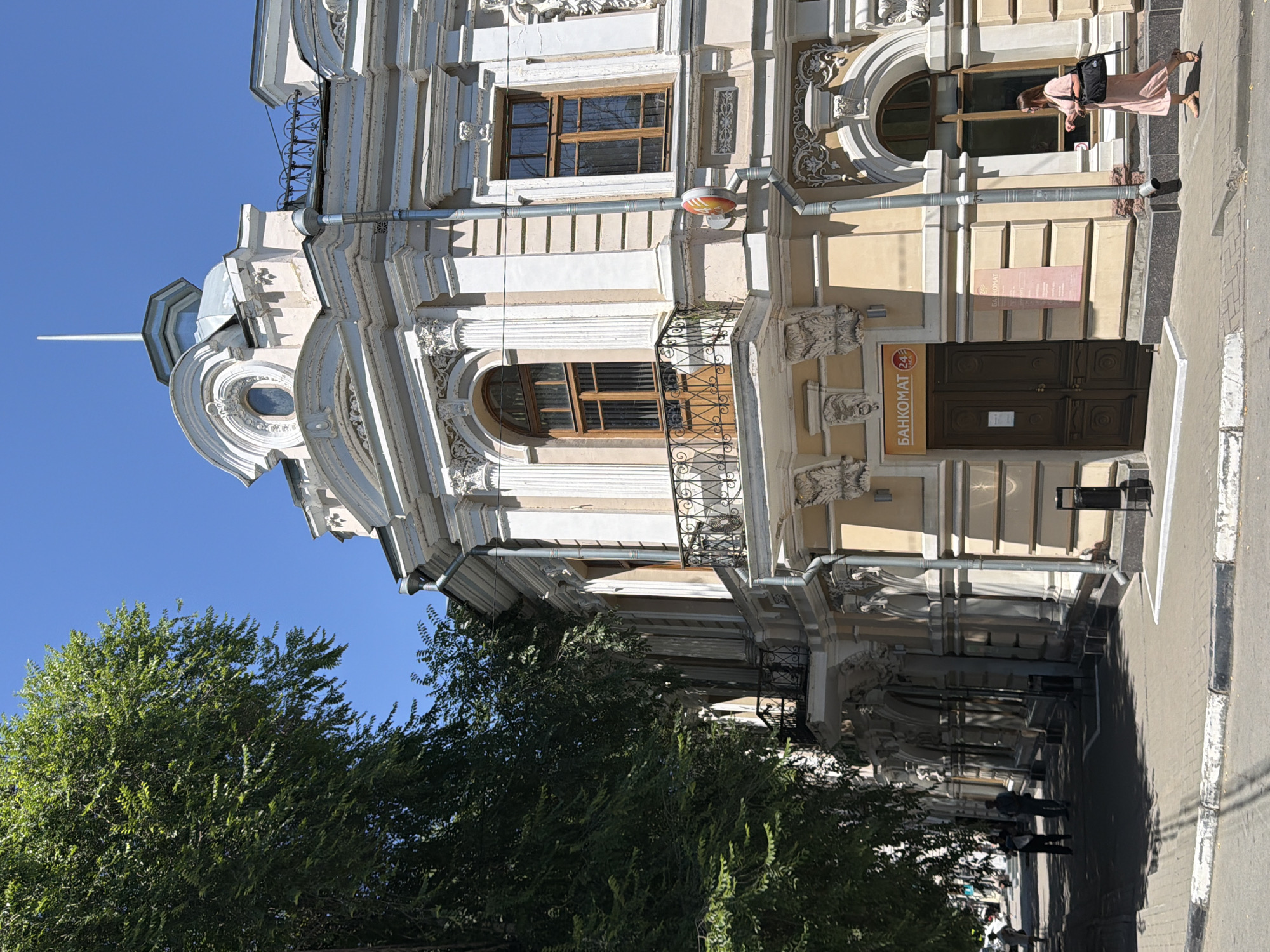 A medium shot captures a street-level view of a light beige, multi-story building with ornate classical architectural details, including columns, decorative reliefs, and a rounded pediment. A large green tree partially obscures the left side of the building, with its branches and leaves reaching towards the bright blue sky. The building's facade features large, arched windows with wooden frames, some with wrought-iron balconies. A prominent orange sign above a double wooden door reads "БАНКОМАТ 24" (ATM 24), indicating a bank or ATM service. A woman in a light pink dress and sandals walks on the sidewalk to the right of the entrance, carrying a black handbag. To the left of the building, people walk along the sidewalk in front of more storefronts, and a few parked cars are visible in the distance. The street is paved with light gray bricks, and a curb separates it from the sidewalk. The scene appears to be a sunny day in an urban environment. This is the building of the former Bank of Moscow, located at 18 Frunze Street in Taganrog, Russia.