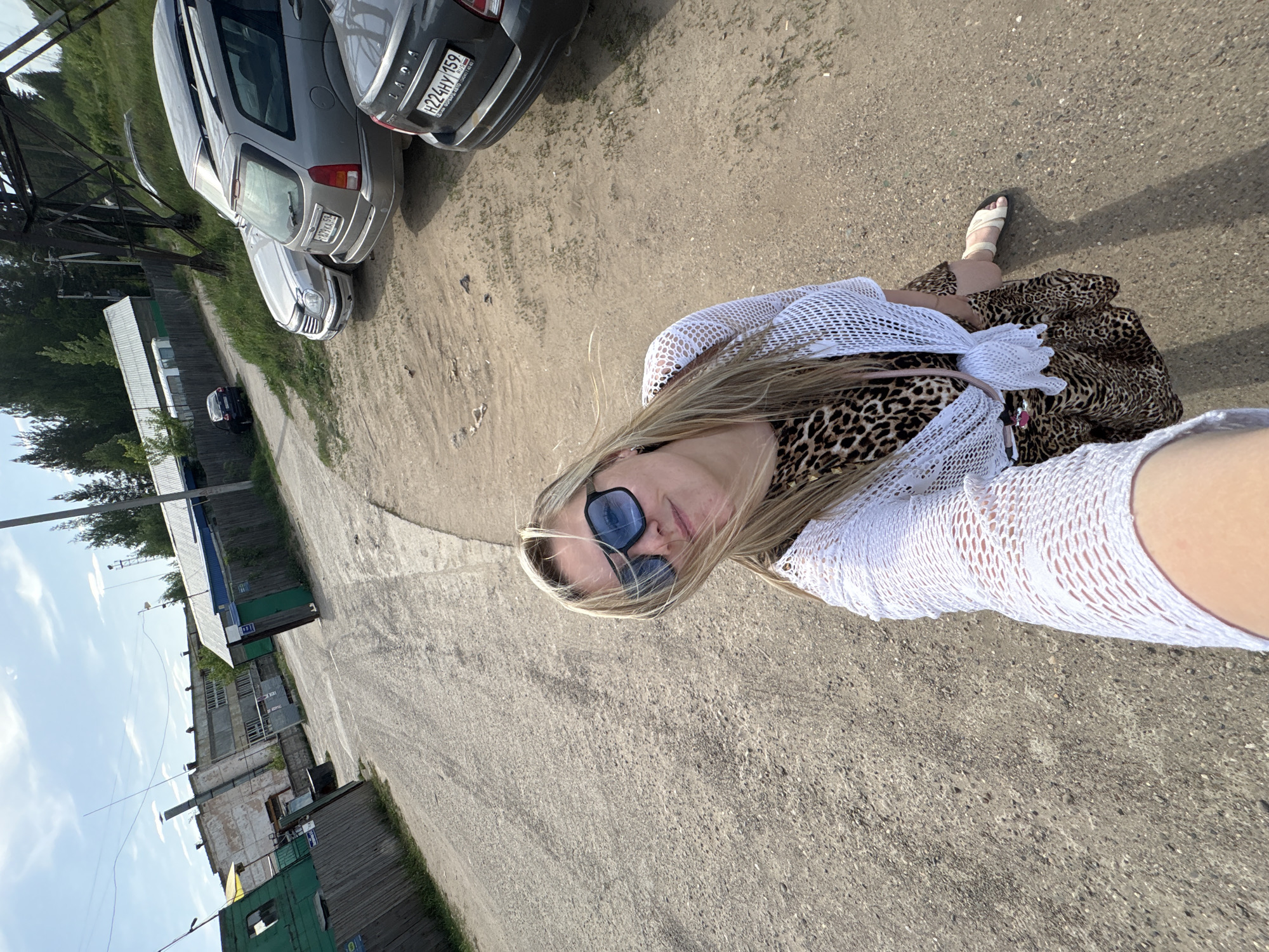 The image is a selfie taken outdoors. A woman wearing sunglasses, a white crochet top, and a leopard-print skirt is partially visible. She is standing on a paved area, likely a road or parking lot. Several parked cars are visible in the background, including a grey Lada with the license plate "H224HY 159" and a silver car with the license plate "Y109XA 59." A building complex with grey and green structures is also visible. Trees and power lines are in the background. The location is likely the city of Noviy, Russia.