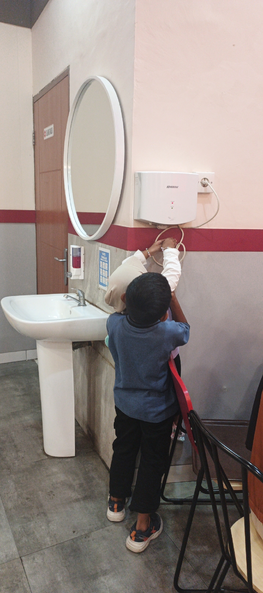 In a public restroom in Surabaya, Indonesia, two young children are interacting with a wall-mounted electric hand dryer. The child on the left, wearing a blue t-shirt and black pants, stands on a red chair, reaching up to the hand dryer with both hands. Their arms are extended, and they appear to be manipulating the cord or the unit itself. To their right, another child, partially obscured, also reaches towards the hand dryer, wearing a beige hijab and white long-sleeved shirt.
The hand dryer is white, with the "KRISBOW" logo visible. It is mounted on a wall with a pink and red striped accent. Below the hand dryer, on the wall, is a blue sign with a grid of icons, likely depicting hand-washing instructions. To the left of the hand dryer, an oval mirror with a white frame is mounted on the wall. Below the mirror is a white pedestal sink with a silver faucet and a wall-mounted soap dispenser. The floor is tiled with gray concrete. A wooden door with a sign on it is visible on the far left. The overall impression is a public facility, possibly a restaurant or shopping mall restroom, where children are curious about the amenities.