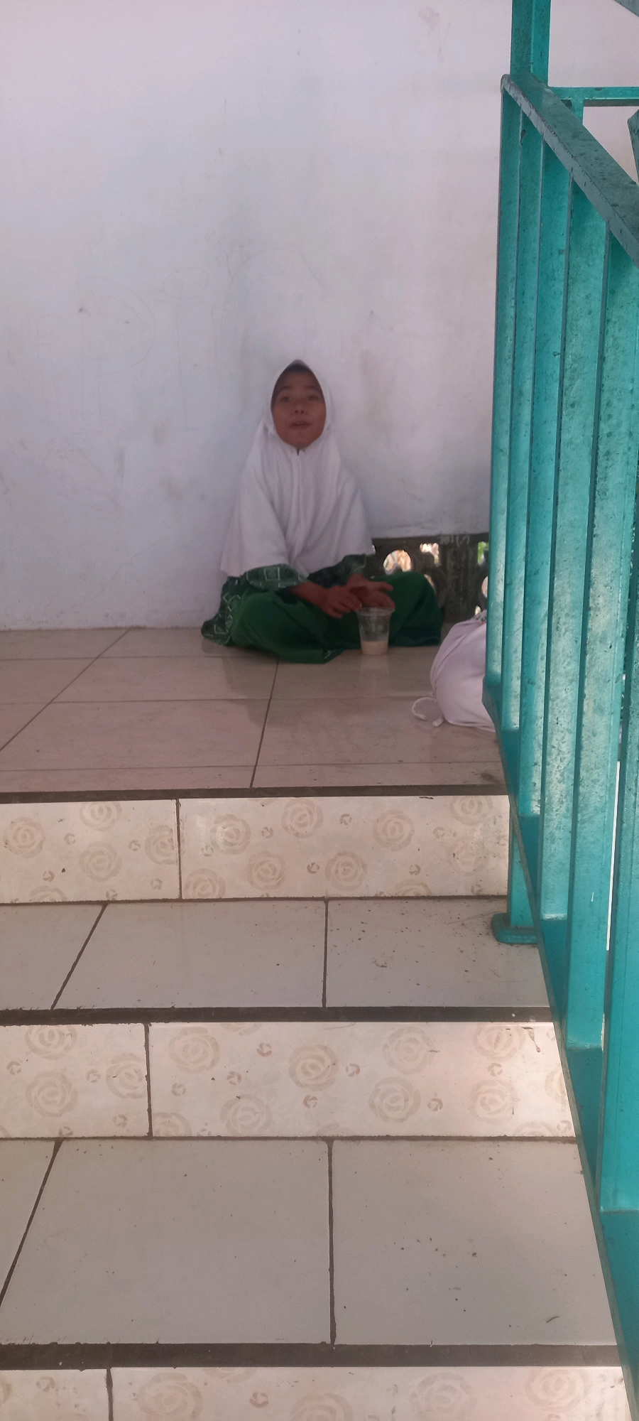 An undated, eye-level, medium-shot photograph captures a young girl sitting cross-legged on a tiled floor, leaning against a white wall. She is wearing a white hijab and a green and white patterned skirt. Her hands are clasped in front of her, and a clear plastic cup containing a light-colored liquid is placed on the floor to her right. To her left, a white bundle rests on the floor. The girl is looking directly at the camera. The floor is tiled with a light-colored base and a rose pattern. In the foreground, a set of stairs with similar tiles is visible, leading down. A bright turquoise railing runs along the right side of the frame. The background wall is textured and appears aged. The lighting suggests an outdoor or semi-outdoor environment. The scene is set in Pontang, Indonesia.