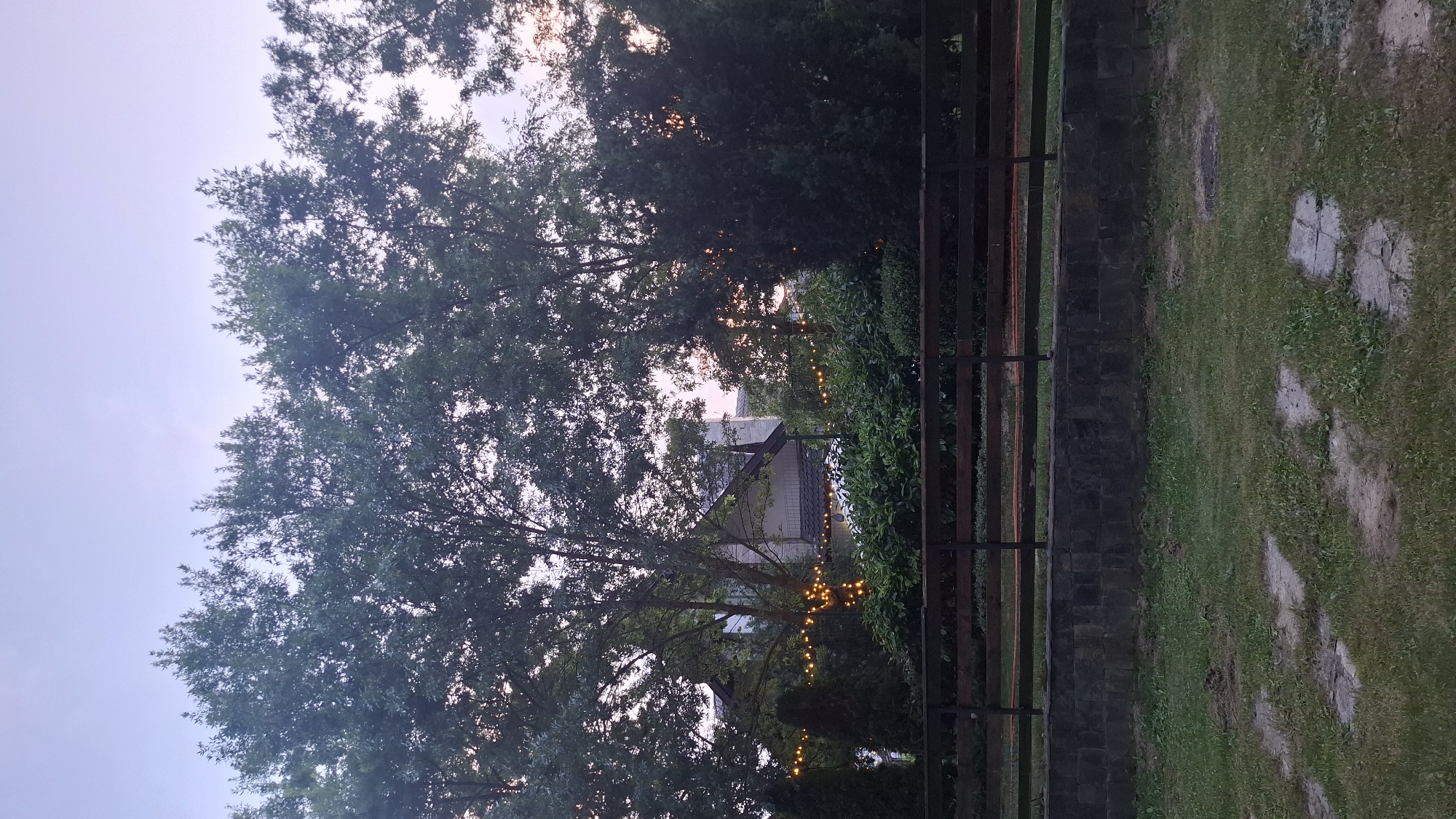 The image depicts an outdoor scene at dusk, likely in a garden or park setting in Razlog, Bulgaria. The main subjects are lush green trees and foliage, with a building visible in the background partially obscured by the vegetation. String lights, emitting a warm yellow glow, are draped across the trees and bushes, suggesting an evening gathering or decorative ambiance. A dark wooden fence runs horizontally across the middle of the frame, separating the garden area from a grassy patch with stone stepping stones on the right. The sky is a soft gradient of pale blue and hints of pink, indicating the twilight hour. The overall mood is serene and inviting, evoking a peaceful end to the day. There is no visible text in the image.