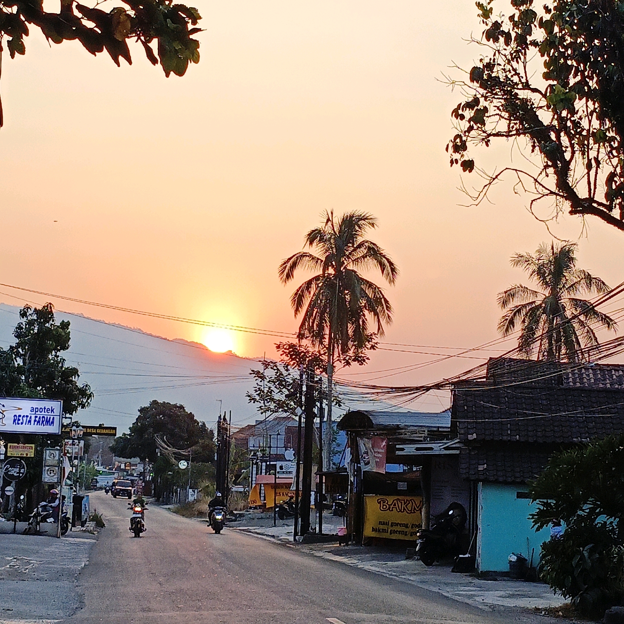 The image depicts a street scene in Gedangan, Indonesia, at sunset. The sky is a vibrant gradient of orange and yellow, with the sun partially obscured by mountains in the distance. Silhouetted palm trees and other vegetation frame the scene. On the left side of the road, a white sign for "apotek RESTA FARMA" is visible, along with other smaller signs and what appear to be storefronts. Several motorcycles are on the road, with some riders visible. One car is also present on the road. The road itself is paved and appears to be a main thoroughfare with buildings and power lines lining its sides. The overall atmosphere is that of a bustling town during the golden hour.