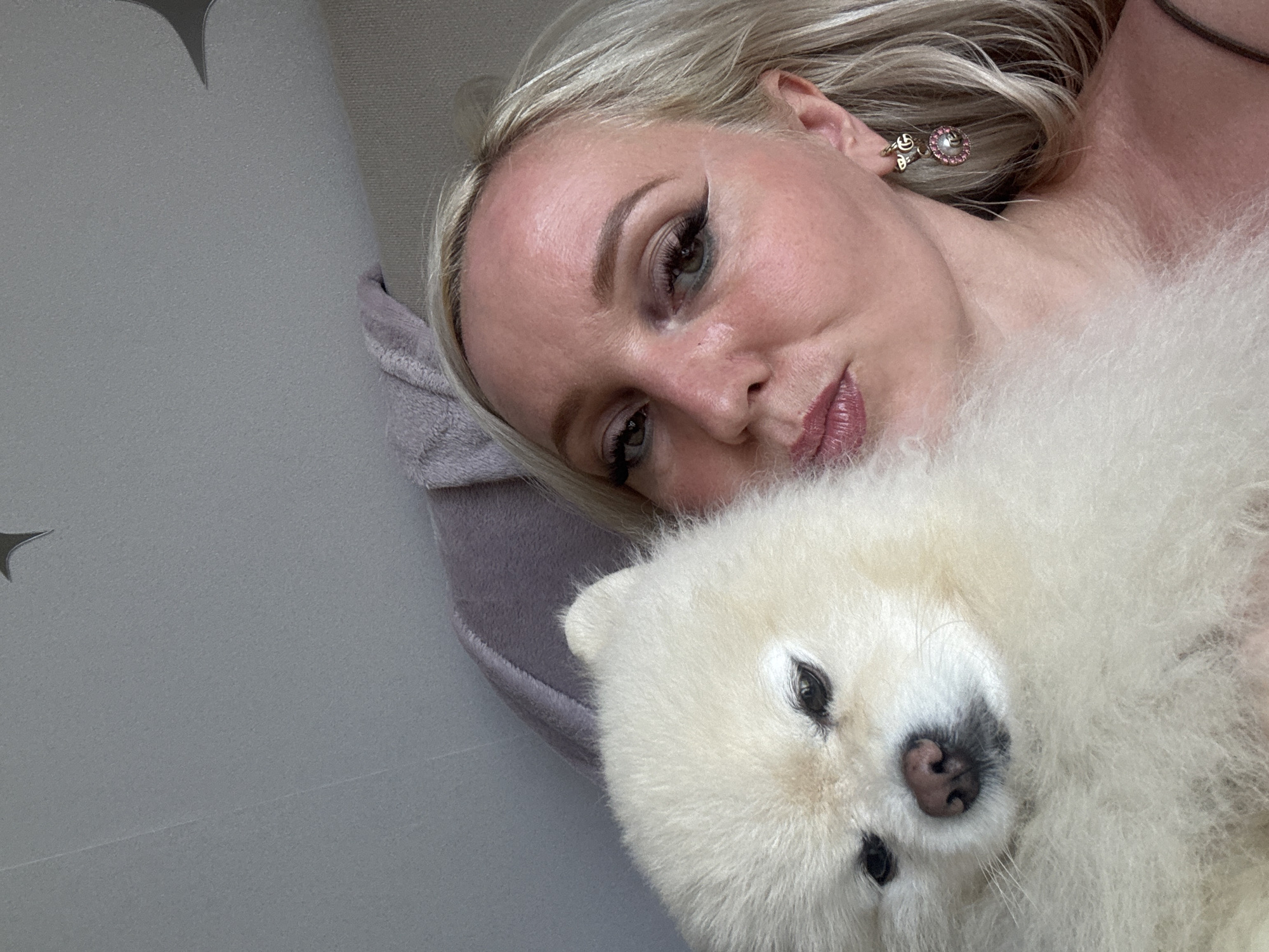 A close-up selfie captures a woman and a fluffy white Pomeranian dog. The woman, with blonde hair and heavy eye makeup, is puckering her lips towards the camera, holding the dog close to her face. The dog's dark eyes and pink nose are visible. The background consists of a light-colored, textured surface, possibly a wall or ceiling, with a faint shadow. The woman is wearing a dark-colored top with a thin strap, and a dangling earring with pink stones and a 'G' logo. The image appears to be taken indoors, in what could be a residential setting. There is no indication of the city of Komsomolsk-on-Amur, Russia.