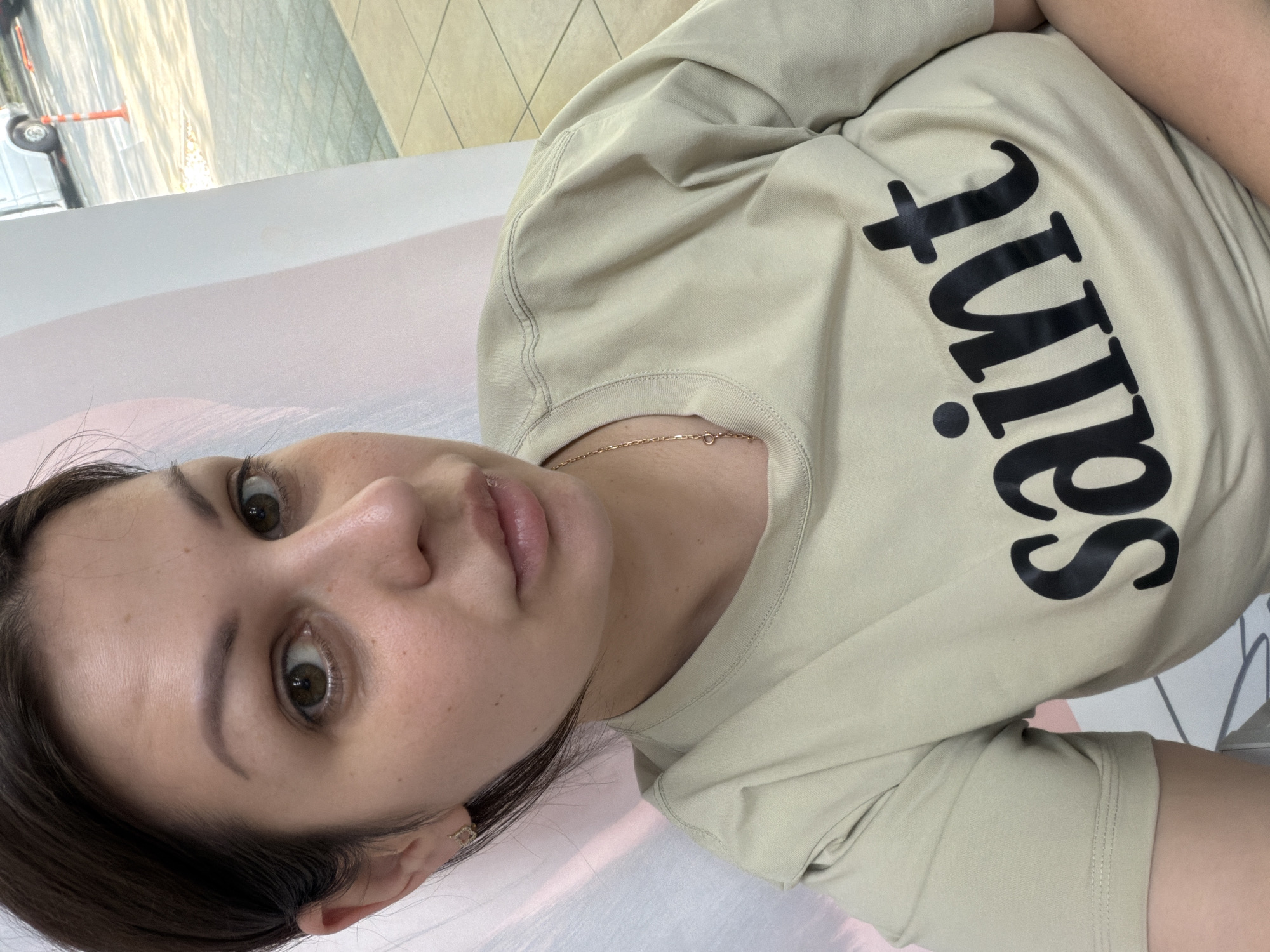 A close-up selfie of a young woman is captured vertically. She is wearing a light beige t-shirt with the word "Saint" printed in large black letters. A delicate gold chain is visible around her neck. The woman has brown hair and expressive green eyes, with natural makeup. Her lips are slightly parted, revealing a subtle gloss. She is wearing a small gold hoop earring in her left ear.
The background is out of focus, but suggests an indoor setting with tiled walls and flooring, possibly a commercial or public space. A hint of an outdoor scene with a barrier and a vehicle is visible in the upper left corner. The overall lighting suggests daytime. The woman's facial expression appears relaxed and direct, looking towards the camera. The image is cropped to focus on her face and upper body.