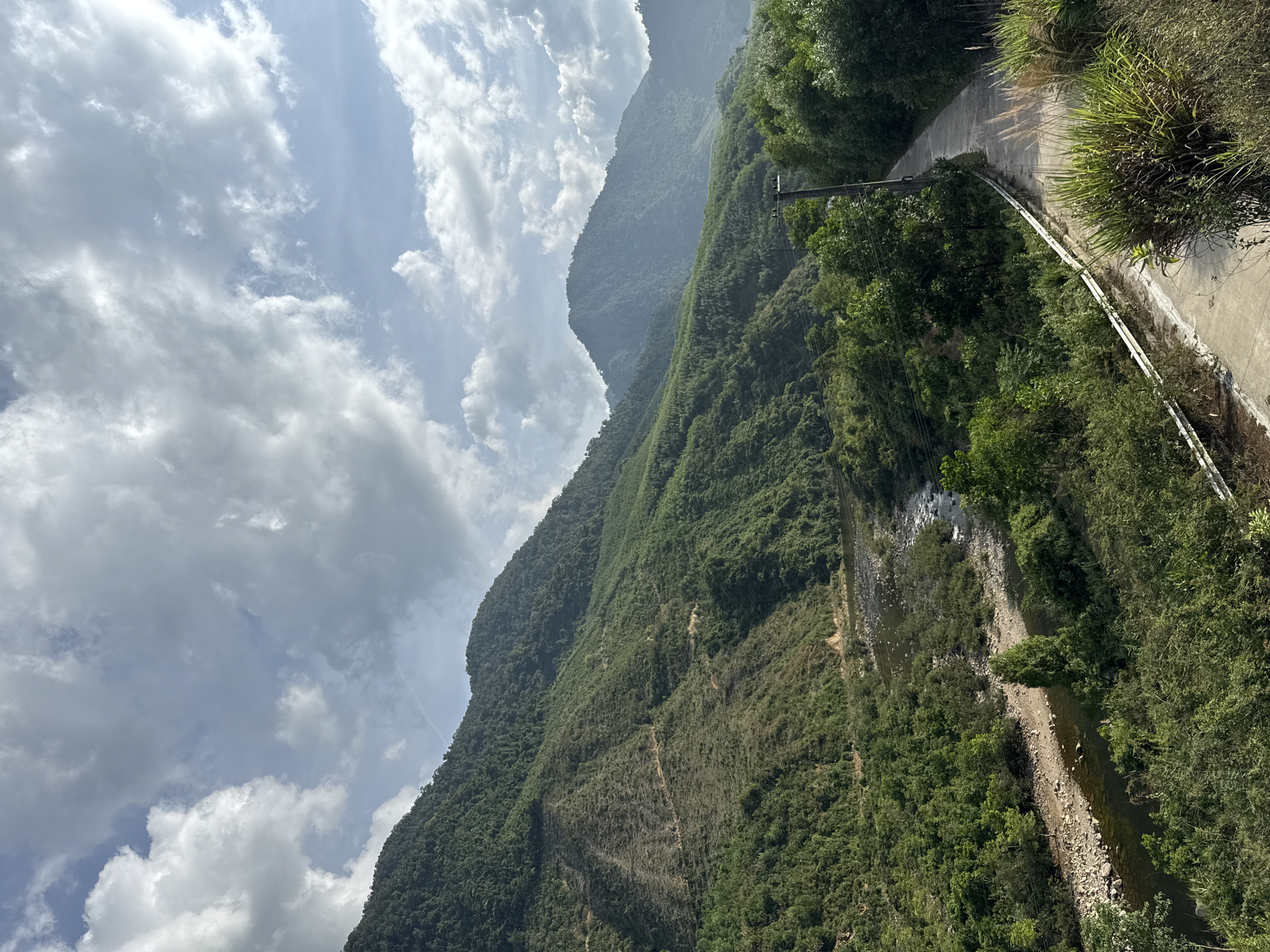 The image depicts a mountainous landscape adjacent to a narrow road. The road, constructed of concrete, runs alongside a steep, heavily vegetated slope leading down to a shallow river. The mountains are densely covered with trees and foliage. The sky is partially obscured by cumulus clouds. The image does not show any people or indicate any specific actions or interactions. There is a power line pole next to the road. There are no clues in the image to indicate that the location is the city of Đà Nẵng, Vietnam.