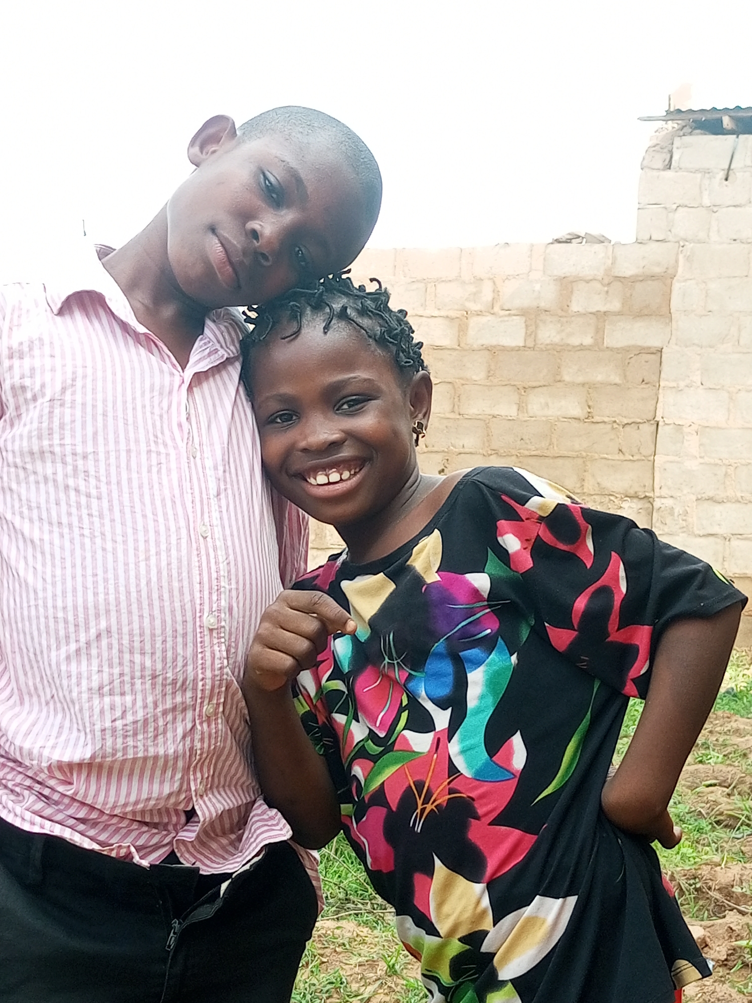 Two children, a boy and a girl, are posing for a photograph outdoors in Tempure, Nigeria. The boy, on the left, is wearing a pink and white striped button-down shirt and black pants. He has a short haircut and is leaning his head towards the girl. The girl, on the right, is wearing a black dress with a colorful floral print. She has her hair braided and is smiling broadly at the camera, pointing towards the viewer with her right hand. Behind them, a light-colored brick wall with some rubble on top is visible, suggesting an unfinished or damaged structure. The ground is covered with dry grass and soil. The overall lighting is bright, indicating an outdoor setting during the day.