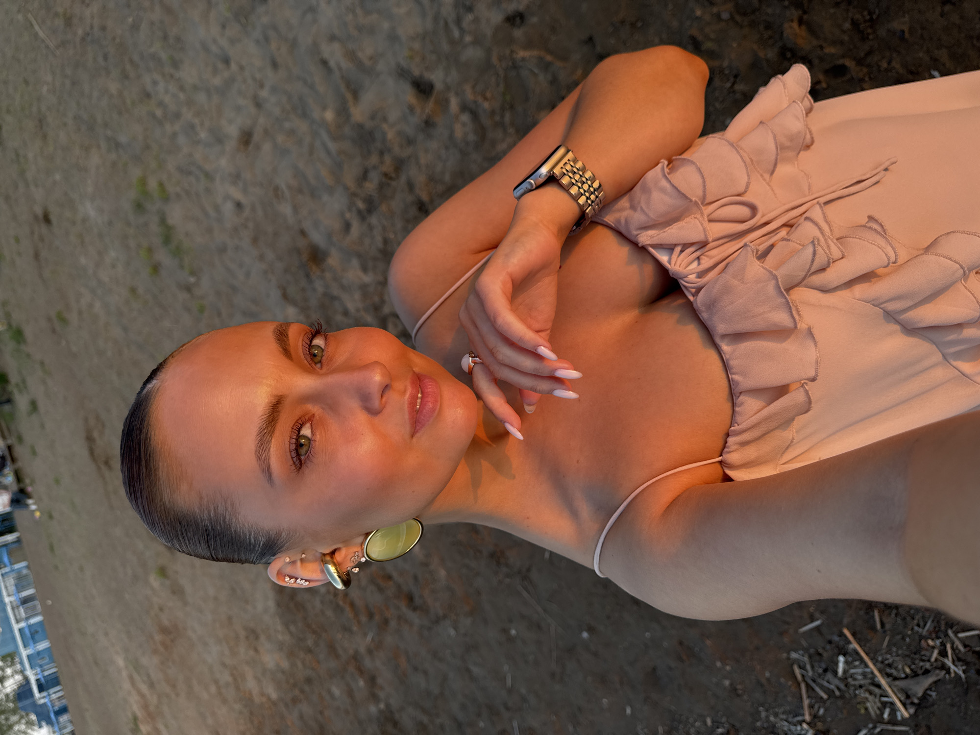 A woman with light brown hair pulled back and light skin is laying on the ground in an outdoor setting. She is wearing a light pink dress with ruffled details and thin straps. She also has several earrings in her left ear, a ring on her finger, and a wristwatch on her left wrist. The ground appears to be a mix of sand and debris. A portion of a building is visible in the bottom left corner.
While the query mentioned the city of Chaikovsky, Russia, this detail cannot be verified solely from the image content. The setting appears to be an outdoor location with some proximity to a building, but specific location details are impossible to confirm from the image alone.