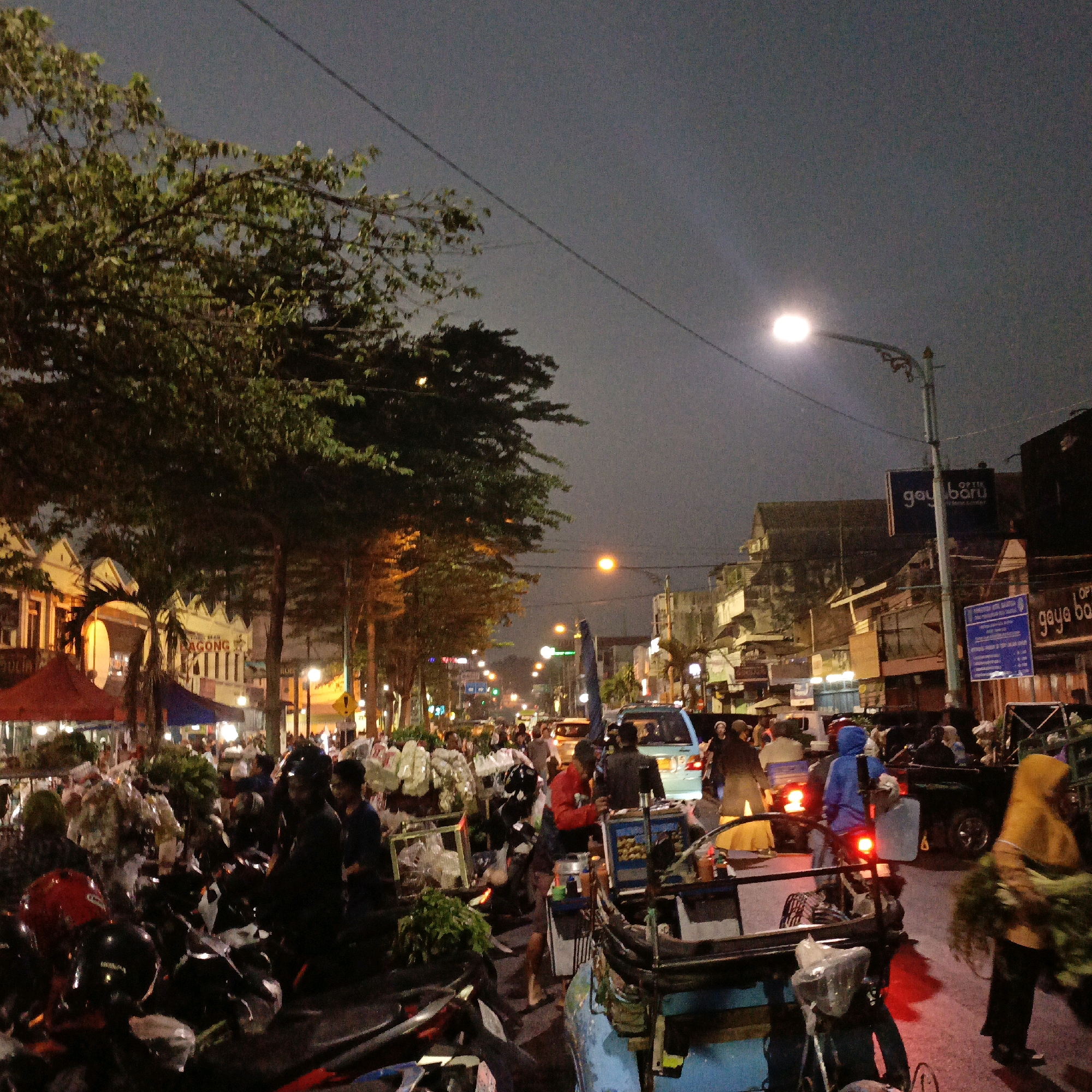 This image captures a bustling night scene in Salatiga, Indonesia, likely along a street lined with shops and market stalls. The sky is dark, suggesting it is late evening or night, and illuminated by streetlights and the lights from buildings and vehicles.
In the foreground, a collection of parked motorcycles, some with helmets on them, indicates a busy area. To the left, several market stalls are visible with red and blue awnings, displaying a variety of goods, including what appears to be produce and flowers. People are gathered around these stalls, browsing or working.
The street itself is filled with vehicles, including cars and what appears to be a bright blue van. Pedestrians are also present, some walking along the sidewalk and others interacting with vendors. The buildings lining the street display signs, one of which reads "OPTIG baru" and another "gaya ba", suggesting commercial establishments. The overall atmosphere is one of lively activity, typical of a night market or a busy urban street at night.