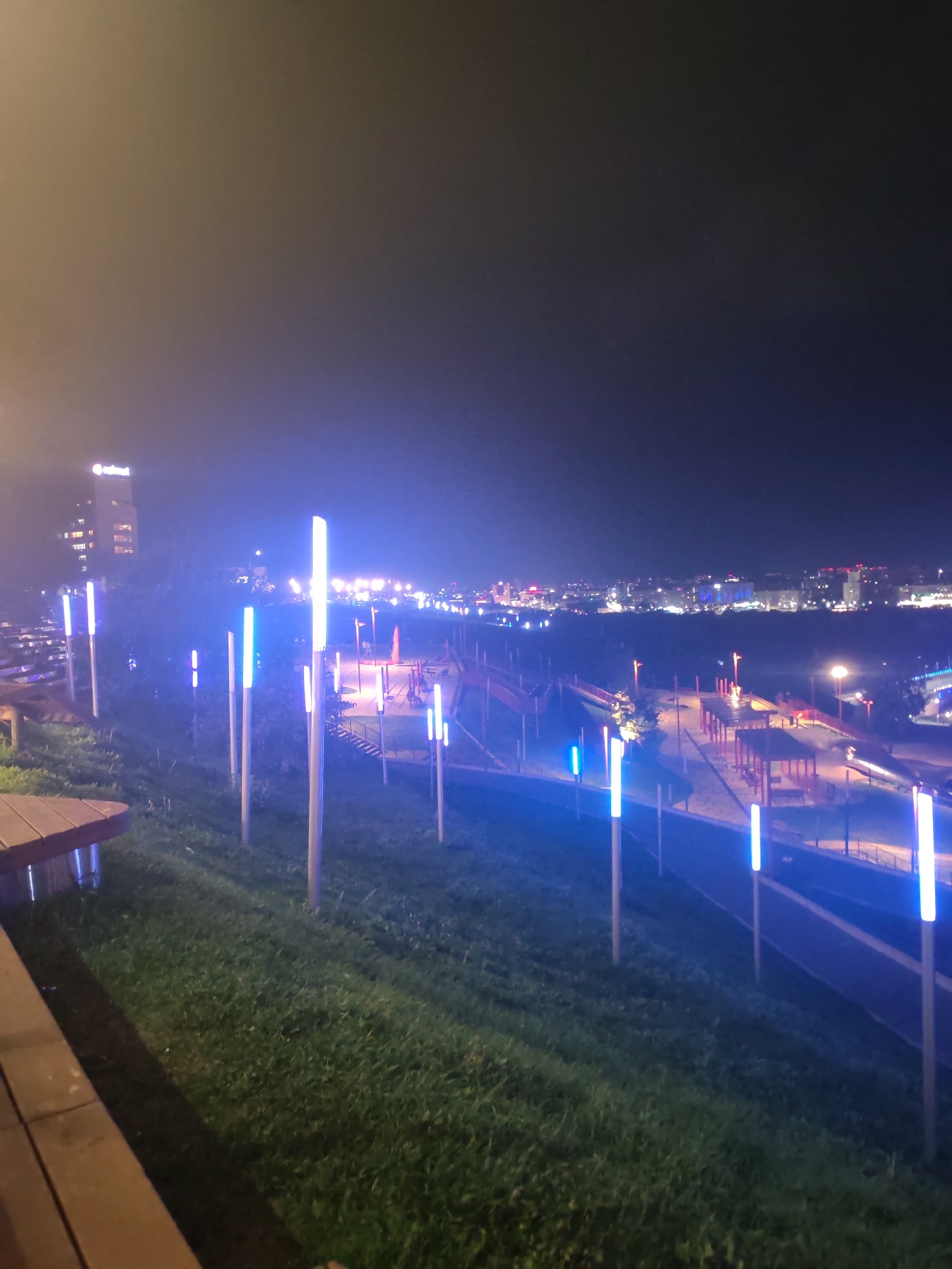 The image captures an urban landscape at night, likely in Nizhny Novgorod, Russia, given the lighting and architectural style. In the foreground, a grassy slope is illuminated by a series of tall, slender lights emitting a bright blue glow. To the left, a wooden bench with metal supports is partially visible, suggesting a public seating area.
Further into the scene, a pathway winds through the illuminated landscape, bordered by more blue lights. Beyond the pathway, a children's playground with red structures is visible, illuminated by a mix of its own lights and the ambient city glow.
In the background, the city skyline is a tapestry of lights. A prominent tall building, possibly an office or residential complex, stands out with its illuminated windows and a logo at the top that reads "GLOBALNET". The rest of the skyline is a blur of lights from various buildings, indicating a densely populated urban area. The overall atmosphere is one of a modern, well-lit city park at night, with a clear view of the cityscape in the distance.
