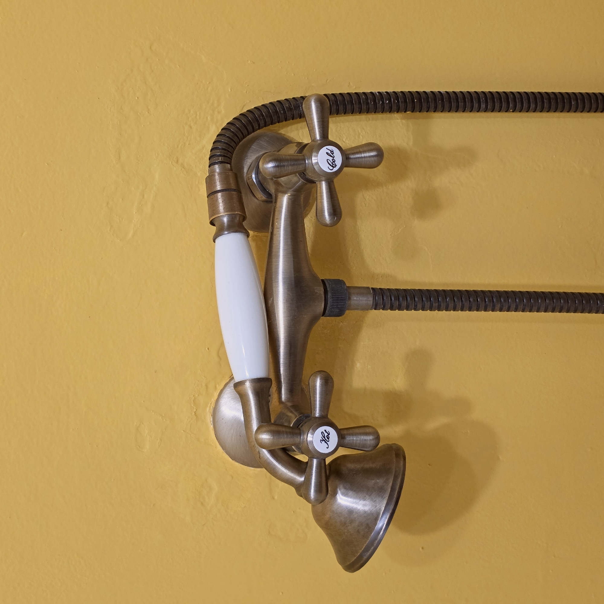 A brass, vintage-style telephone shower fixture is mounted on a yellow wall. The fixture features two cross-handled knobs labeled "Hot" and "Cold", a white porcelain handle, and a flexible metal hose extending horizontally across the wall. The scene lacks identifiable people or actions, focusing solely on the static presentation of the shower fixture and its immediate surroundings. The location, Тэльмінскі сельскі Савет, Belarus, is metadata not visually discernible from the image itself.