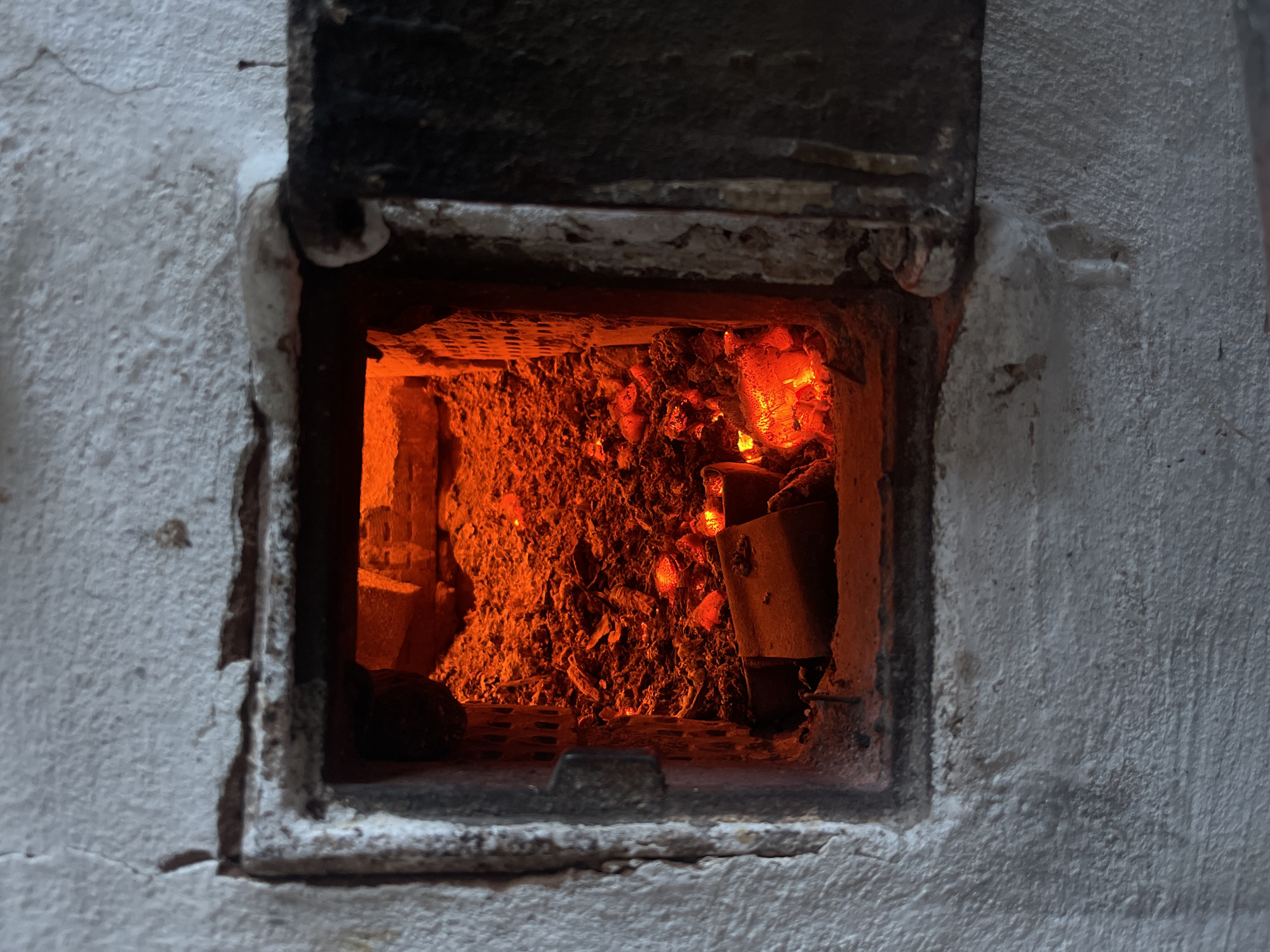 A slightly open door reveals the interior of a white brick oven. Within, glowing embers and charred remains provide the only illumination. No people or subjects are visible. The location is not discernible from the image alone.