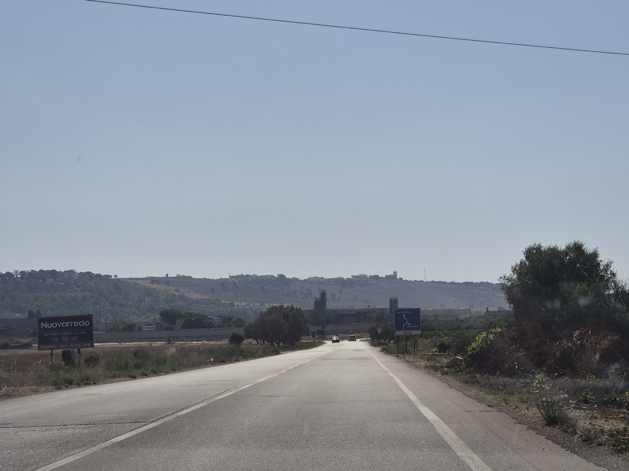 A paved road curves towards the horizon under a clear, light blue sky. The road is bordered by dry, grassy fields and sparse vegetation. In the mid-ground, a billboard reads "Nuovarredo Da Noi ti senti a casa." Further in the distance, a small town with buildings and what appear to be industrial structures sits on a hill. Several vehicles are visible on the road. The scene is likely a rural or semi-rural area.