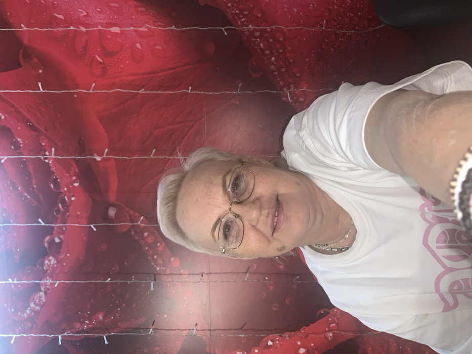 The scene depicts an adult female in an indoor environment within the city of Chaikovsky, Russia. The subject, with fair complexion and short, light blonde hair styled away from her face, wears gold-rimmed eyeglasses and a white, short-sleeved t-shirt featuring a partially visible light pink stylized design on the front. She presents a faint smile while looking directly into the camera. Her right arm is extended into the lower foreground, revealing a patterned bracelet on her wrist. The background consists of a large red panel or wallpaper displaying a detailed print of red rose petals adorned with water droplets. Several vertical strings of small, illuminated white LED lights are suspended in front of this background. A dark, out-of-focus object occupies the extreme lower right portion of the frame.