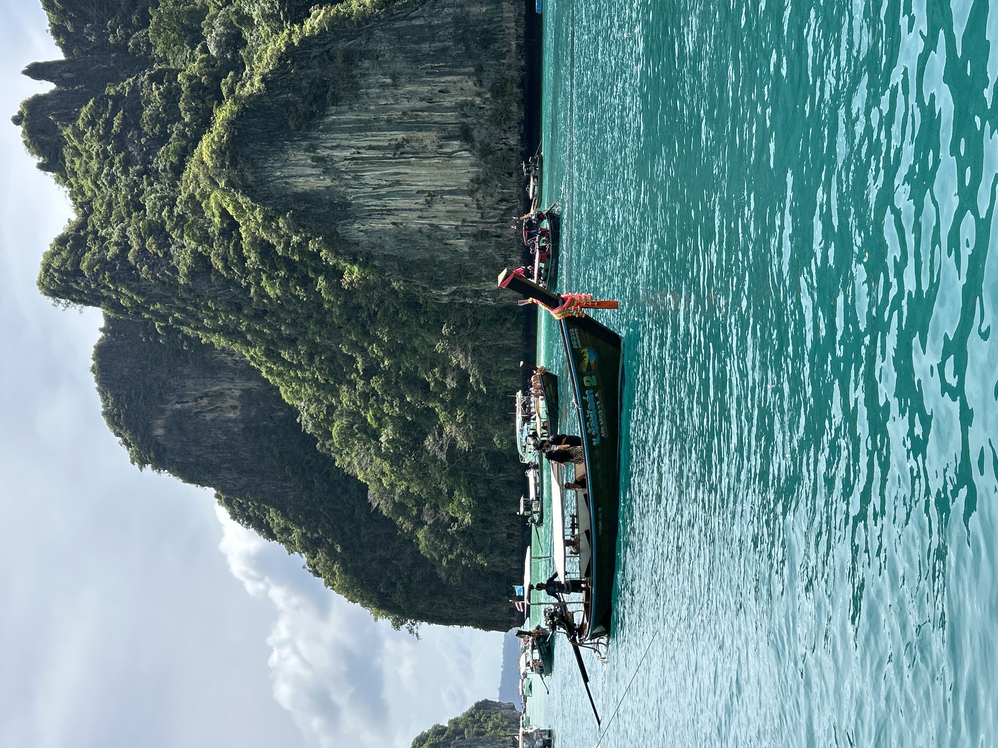 This image captures a serene view of the turquoise waters surrounding Ao Nang, Thailand, with towering limestone karsts covered in lush greenery dominating the background. Several longtail boats, a quintessential mode of transport in the region, are visible on the water. In the foreground, a dark green longtail boat with a white canopy carries a few people. A man stands near the back of the boat, seemingly operating it, while two individuals sit beneath the canopy. Another longtail boat is positioned further back, with several passengers on board. The water exhibits a gentle ripple, reflecting the overcast sky. The overall scene conveys a sense of tranquility and tropical beauty characteristic of Ao Nang.