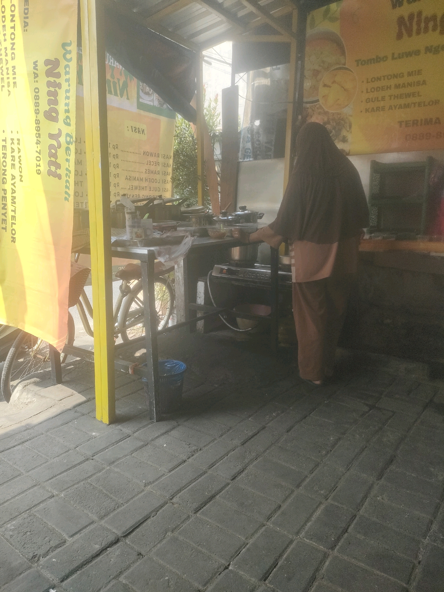 A street-level exterior shot captures a food stall in Surabaya, Indonesia. A woman wearing a dark hijab and a light brown headscarf, with her face obscured, is standing in front of a cooking station. She is wearing a dark hijab and a light brown shirt with matching pants. The stall features a yellow banner with Indonesian text, including "Warung Nasi Tati" and a phone number, "WA: 0889-8954-7019". Behind her, a menu board displays food items and prices in Indonesian. The foreground is paved with dark gray bricks. A bicycle with a basket is parked near the stall. The overall lighting suggests it is daytime.