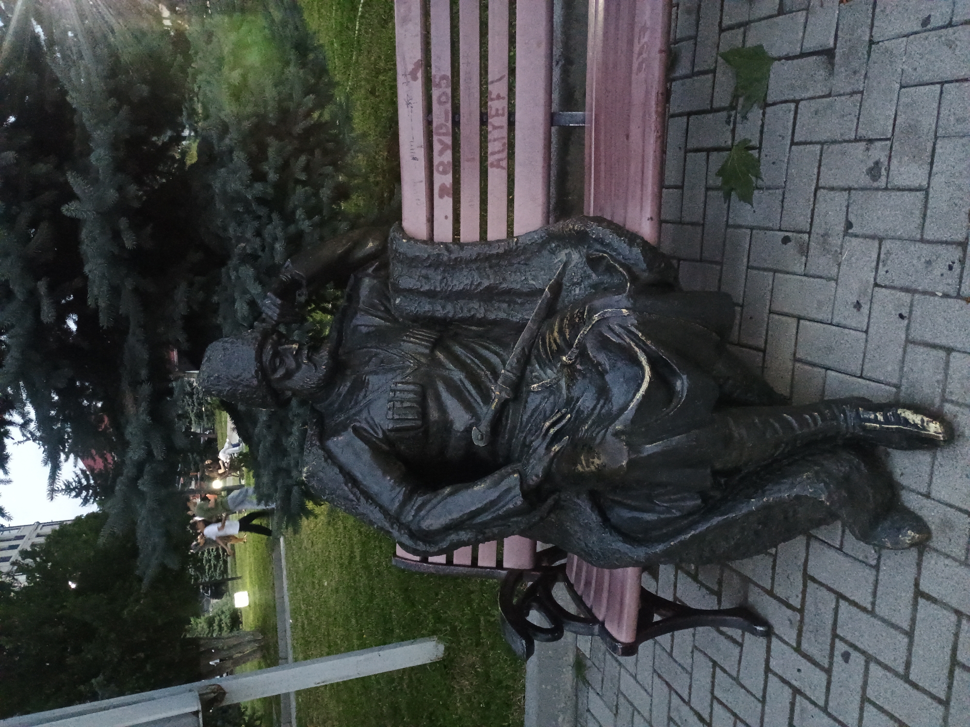 A bronze statue of a man in traditional attire, including a hat, long coat, and boots, sits on a park bench in Gusar, Azerbaijan. The man holds a sword in his lap and has his right arm resting on the back of the bench. The bench is wooden with a metal frame and has graffiti on its slats. The statue is located on a paved walkway in a park with grass and trees visible in the background. There are a few people in the background walking and standing. The lighting suggests it is dusk or early morning.
