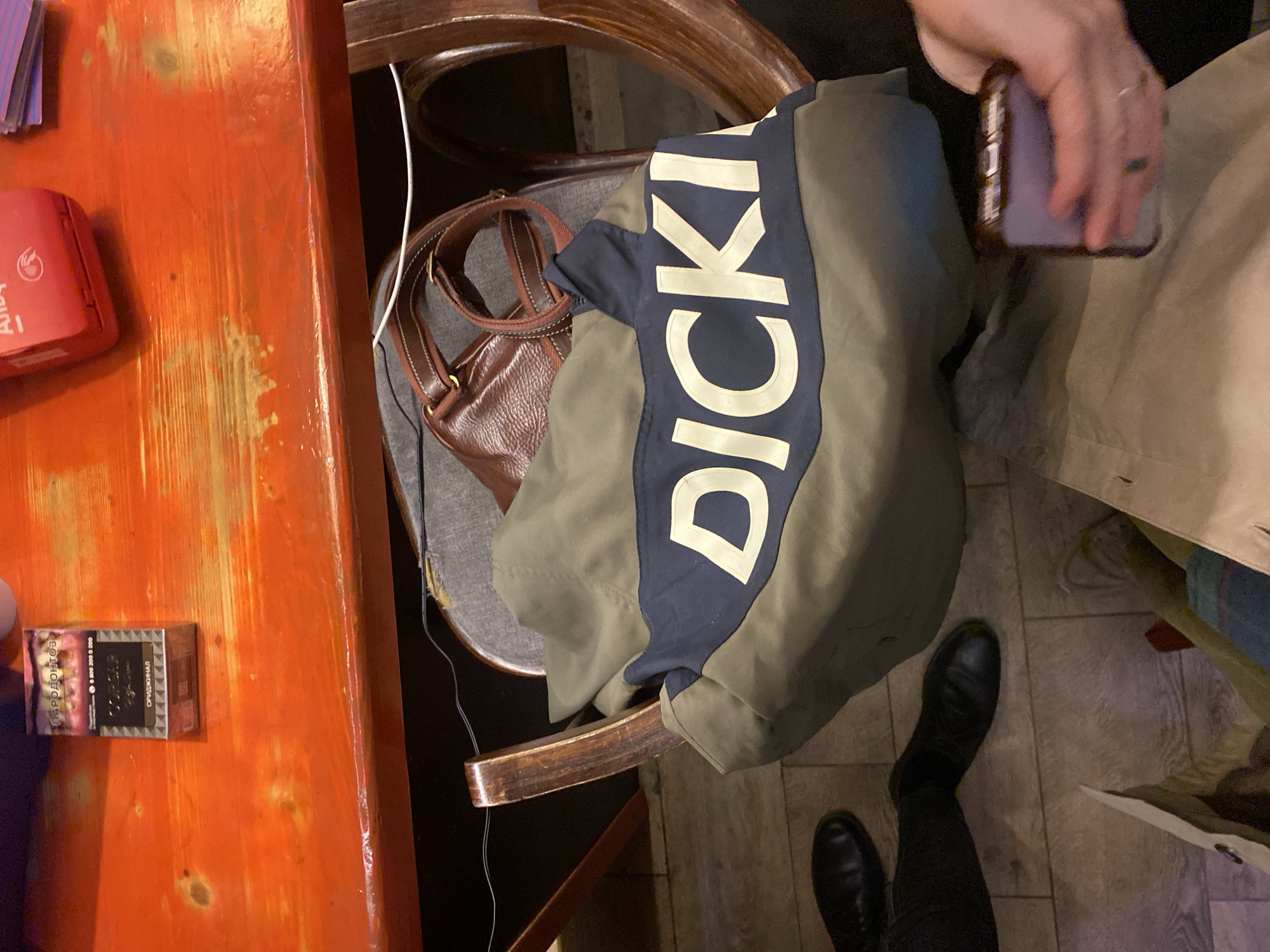 The scene depicts a person holding a smartphone, partially visible, wearing a khaki trench coat and black leather shoes. The person is situated near a wooden chair, which has a brown leather handbag and a folded Dickies jacket on it. The chair is placed next to an orange-colored table with a red box, possibly a cardholder, and a pack of Russian cigarettes called "PARODONTOZ". The environment suggests an indoor location, possibly a restaurant or cafe, with a tiled floor and a mix of natural and artificial lighting. There is no definitive evidence to confirm the location as Moscow, Russia; however, the presence of the Russian brand cigarette suggests a Russian locale.