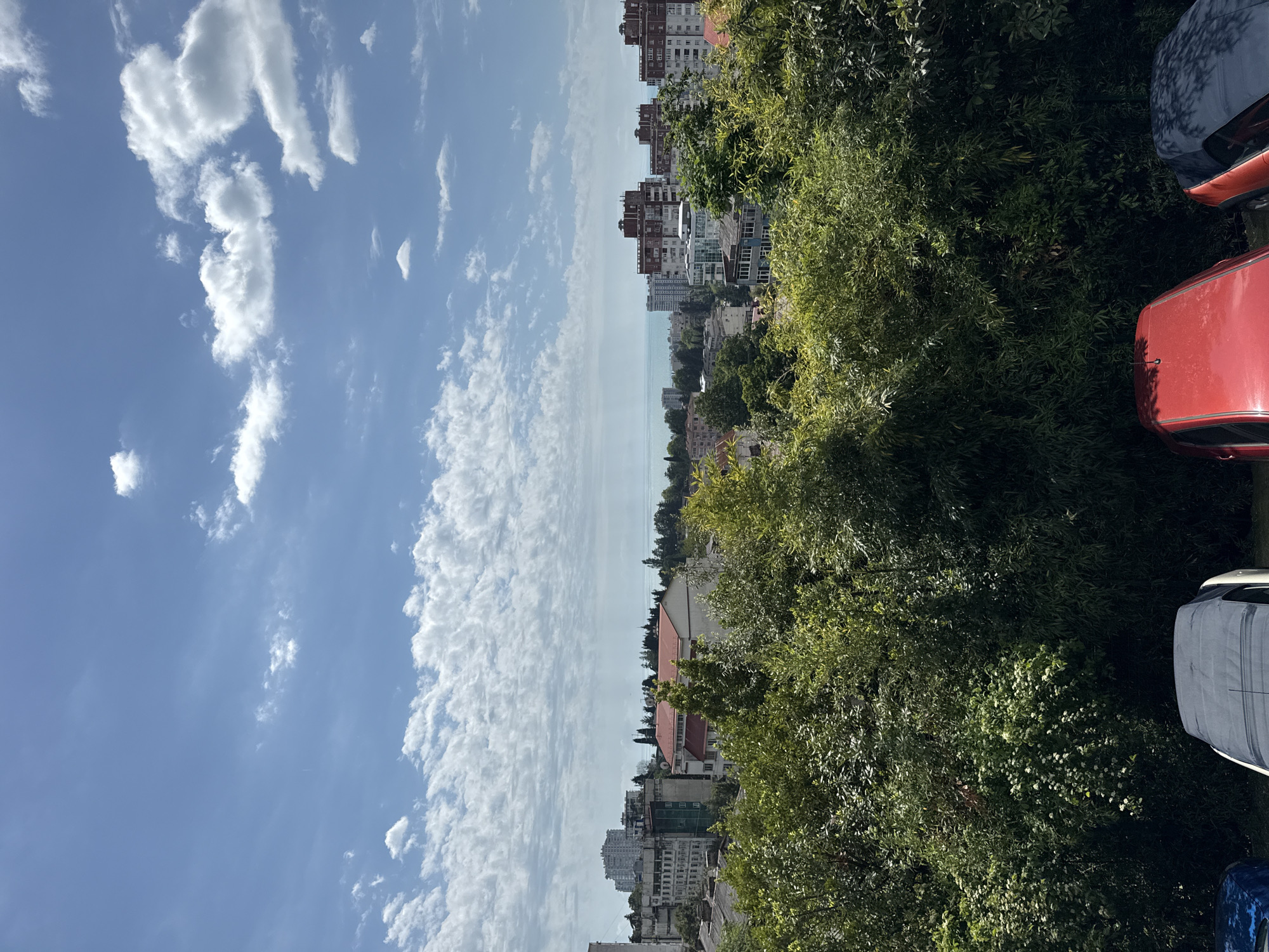 The image shows a view from an elevated position in Sochi, Russia. On the left is a blue sky with scattered white clouds. In the center, there is a cityscape with buildings of varying heights and architectural styles, extending towards the sea. The sea is visible as a thin strip in the distance. On the right, lush green trees partially obscure the view of the city. In the bottom right corner, parts of cars are visible, suggesting a parking area.