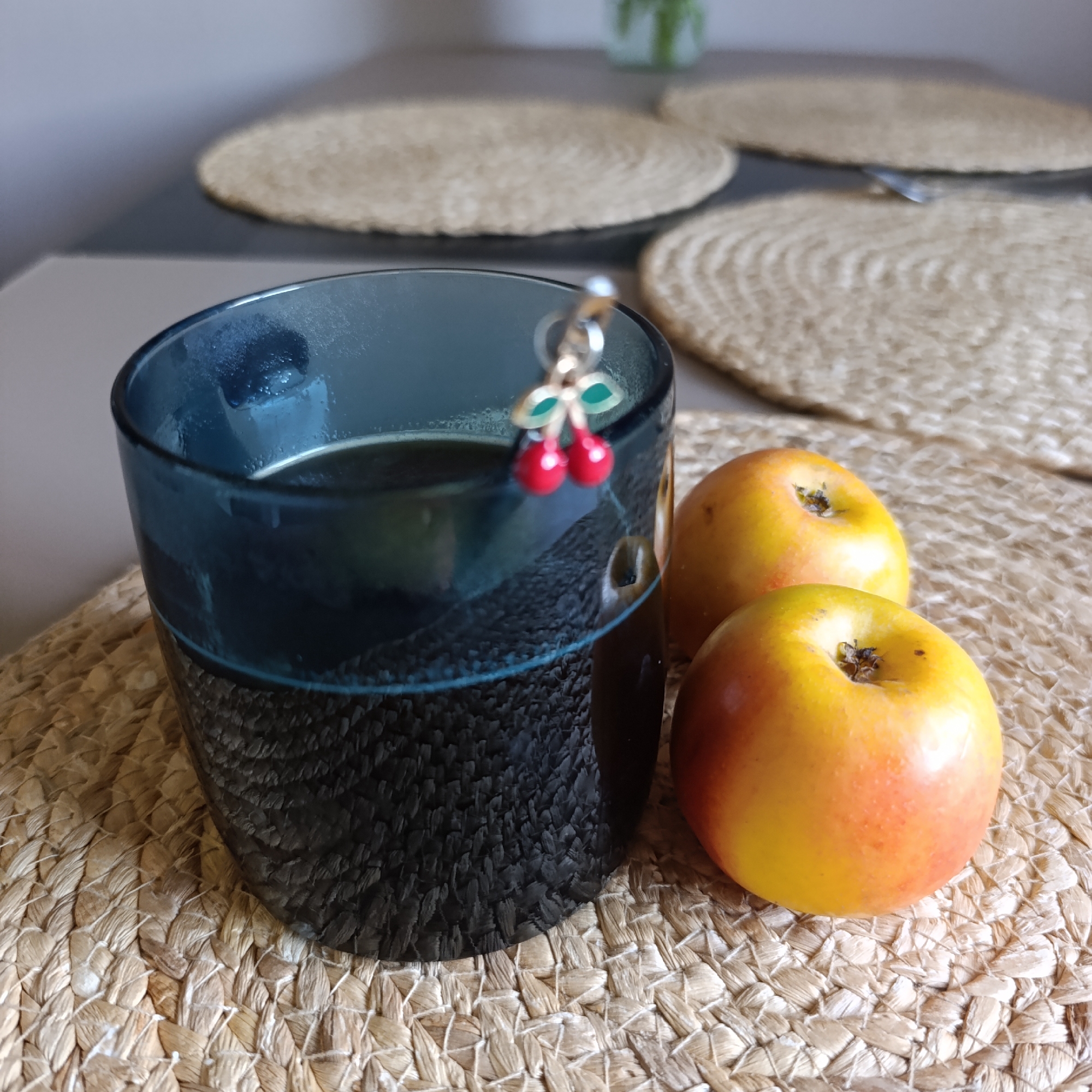 A close-up shot features a dark blue textured glass filled with a dark liquid, likely coffee or tea. Two apples, predominantly yellow with red tinges, sit next to the glass on a woven straw placemat. Additional straw placemats are visible in the blurred background, resting on a two-toned table with a light grey surface and a darker grey lower section. The scene is indoors, with a neutral-colored wall in the background. The cherry-shaped decoration is dangling on the edge of the glass. The location is specified as Марковский, Russia.