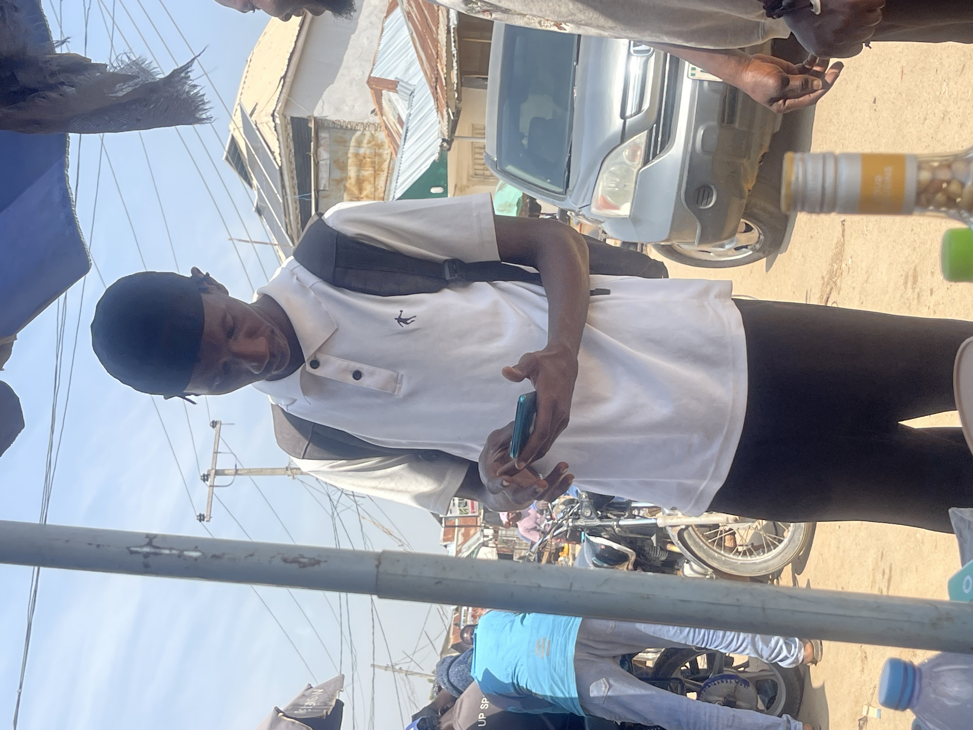 The image depicts a scene in Abuja, Nigeria, likely outdoors with a bright sky suggesting daytime. The primary subject is a young man wearing a black skull cap and a white polo shirt with a small black logo on the chest. He is looking down at a smartphone he holds in his hands. A black backpack is slung over his shoulders. He appears focused on his phone.
In the background, there is a silver minivan or SUV. Further in the distance, structures with corrugated metal roofs are visible, hinting at a semi-urban or market-like environment. There are power lines and poles, common in Nigerian streetscapes. Part of a motorcycle is visible in the lower right quadrant of the image.
The overall impression is a casual, everyday moment captured on the street. The presence of the minivan and the general ambiance suggest a typical day in a bustling area. No specific event is clearly indicated, but the collection of people and vehicles suggests a public space. The focus on the individual's interaction with their phone is a common modern-day activity. There is no discernible text in the image.