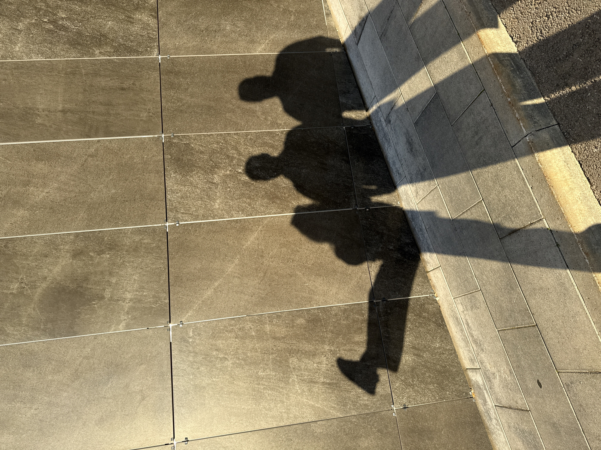 The image depicts the shadows of three individuals cast upon a textured, tiled wall and a paved sidewalk. The wall appears to be made of large, rectangular tiles with a rough, stone-like finish and is divided by thin, vertical white lines. The sidewalk is made of rectangular gray pavers with thin, light-colored grout lines. The lighting suggests it is either early morning or late afternoon, as evidenced by the long shadows. One shadow on the left is elongated and appears to belong to a person with their leg extended. Two more distinct shadows are to the right, suggesting two people standing closer together. The environment is urban, likely a public space. The specific location in Kazan, Russia, cannot be determined from the visual information provided.