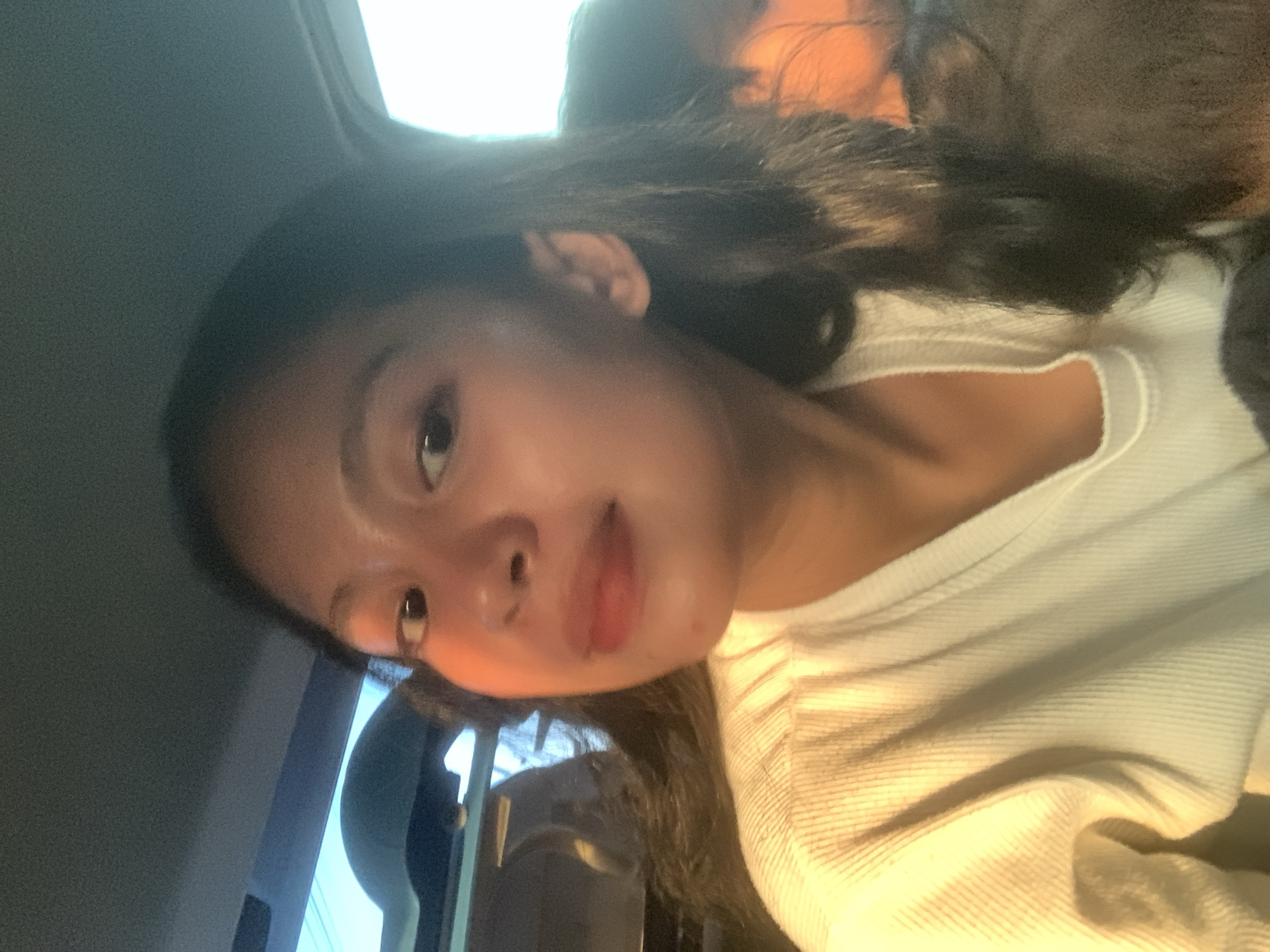 A selfie of a young woman with long, dark hair wearing a white crew neck shirt. She is seated in a car, and the scene appears to be lit with natural light. The location is possibly Batu Batu, Indonesia, although there are no specific details visible in the image to definitively confirm the location.