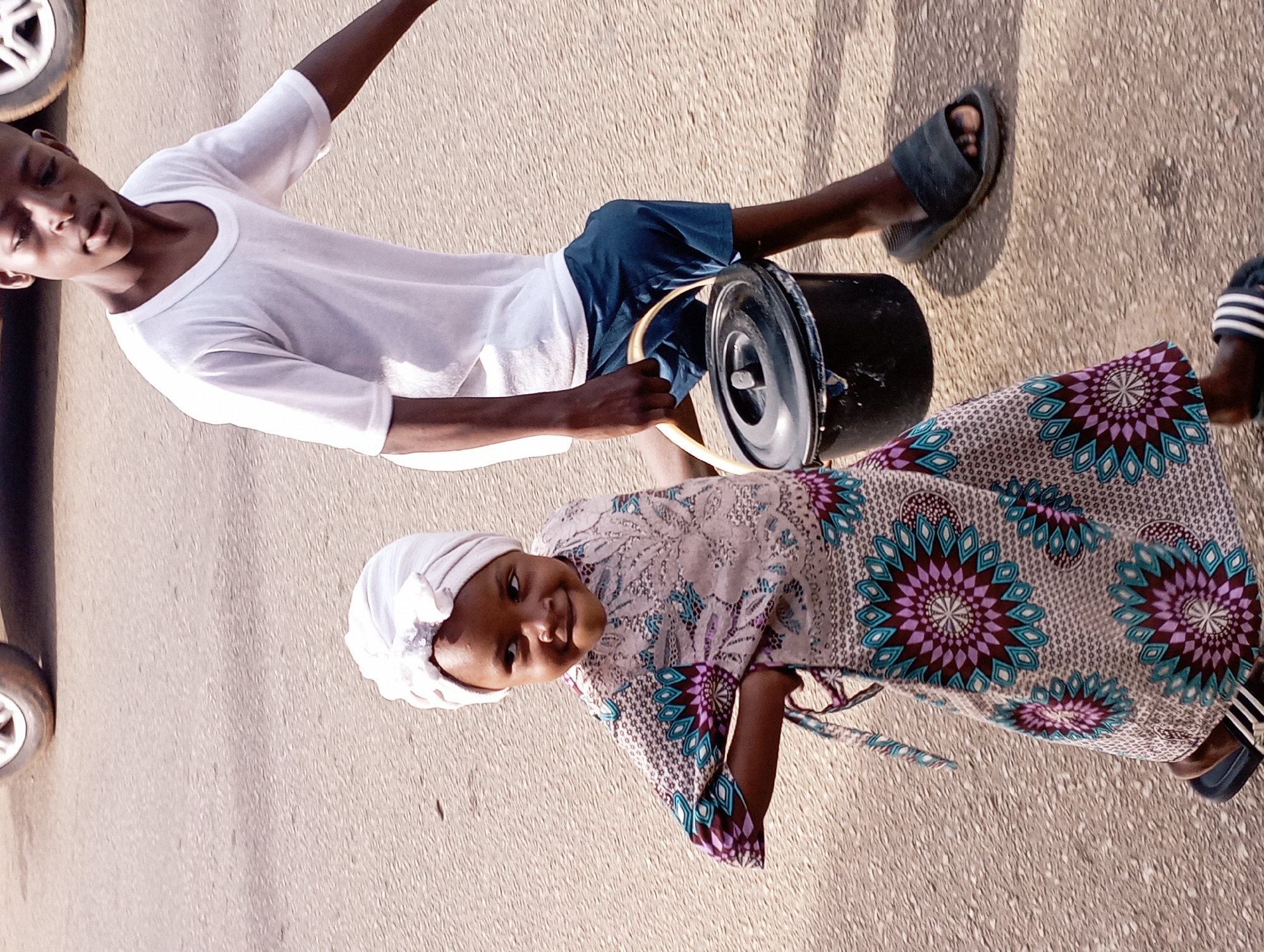 In Jos, Nigeria, on a paved road, a young boy and girl are walking. The boy, on the right, wears a white t-shirt and blue shorts, carrying a black bucket with a yellow handle. The girl, on the left, is dressed in a white headwrap and a colorful patterned dress. Her left hand is on her hip, and she is looking to her left. The image captures them in motion, suggesting they are on an errand. In the background, a portion of a car's wheel is visible. The lighting suggests it is daytime.