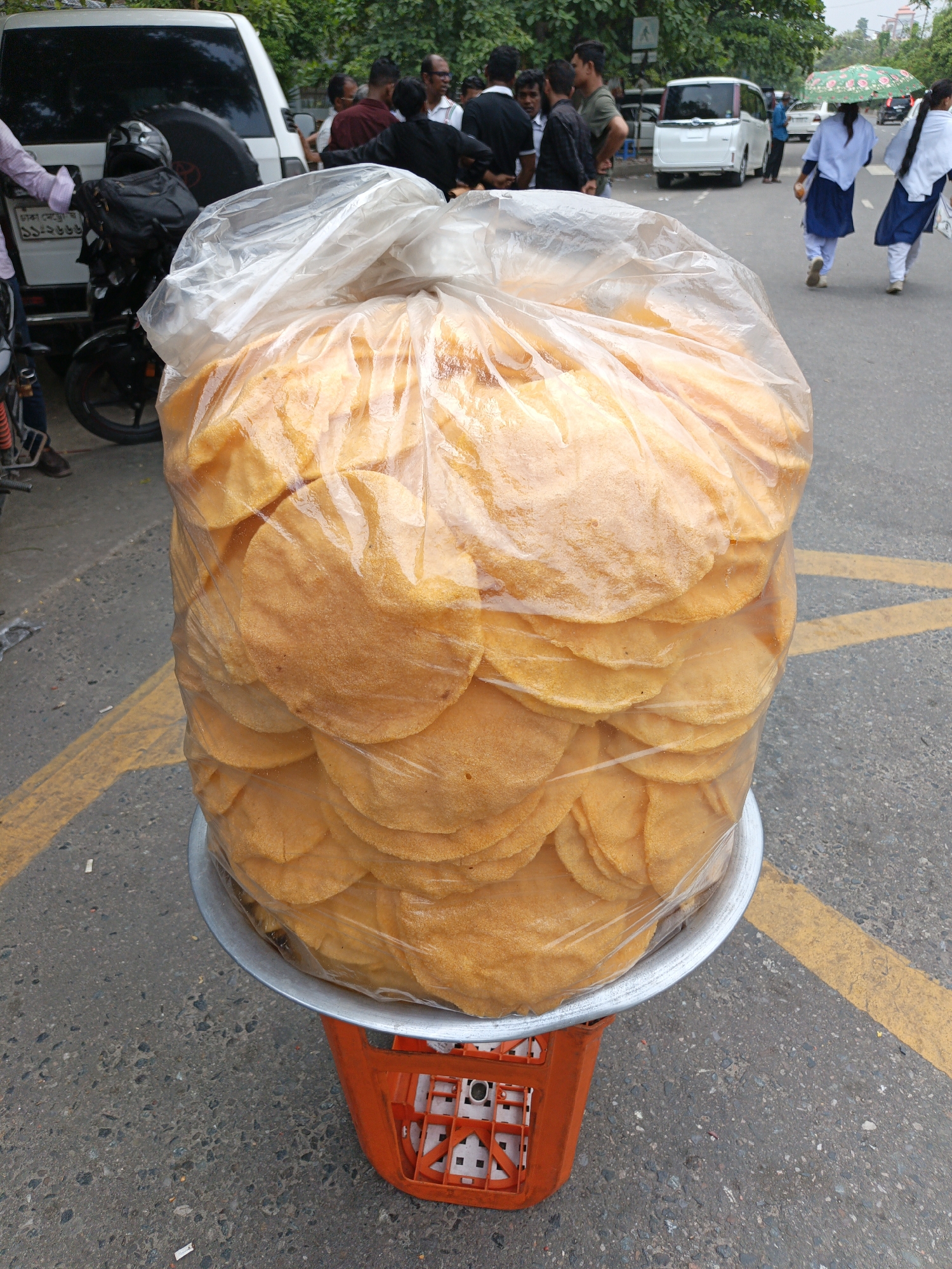 The image shows a large bag of what appears to be flatbread or crispy crackers, displayed on a metal tray which sits on top of an orange plastic crate. The bag is clear plastic and tightly packed with numerous round, golden-brown items. The setup is on an asphalt road with yellow road markings. In the background, there are several people and vehicles, including a white SUV and a light-colored van. Some people are walking on the side of the road, while others are gathered near the SUV. A motorcycle is parked near the SUV. The general setting suggests a street scene in a bustling city.