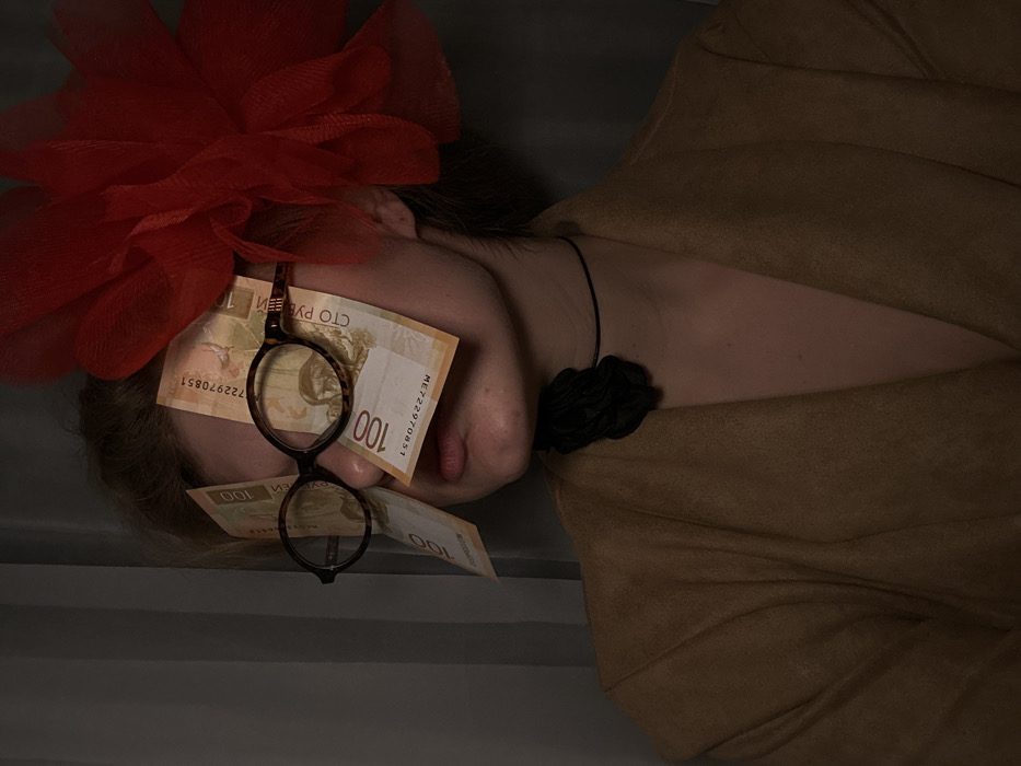In an interior setting in Kazan, Russia, a person is depicted from the upper chest, slightly turned right, facing forward under dim lighting. A large, ruffled, red translucent fabric ornament is attached to the right side of the head. The subject wears round, tortoiseshell-patterned glasses, into which four 100 Russian Ruble banknotes are tucked, obscuring the eyes. The lips are slightly parted. Around the neck is a thin black choker with an irregularly shaped, dark, textured object attached to the front. The person wears a light brown or tan garment with visible lapels. The background is dark and indistinct, with faint vertical lines visible on the left side.