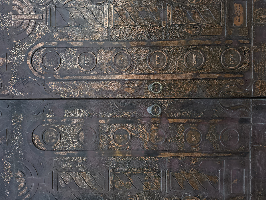 A pair of ornate, dark-patinated metal doors, likely bronze or heavily oxidized copper, is depicted in a full, frontal, close-up view. The doors, divided into two equal vertical panels, exhibit a prominent textured surface with raised relief designs.
Each door panel features a seven-branched menorah with stylized flames at the top section, positioned above a spiraling, fluted column design. Between these and flanking structures, a granular, pebble-like texture covers the surface.
A central vertical band on each door panel contains multiple circular medallions, each embossed with Hebrew text. On the left door, visible inscriptions include "דן" (Dan), "גד" (Gad), "אשר" (Asher), and "יוסף" (Joseph). On the right door, visible inscriptions include "ראובן" (Reuben), "שמעון" (Simeon), "לוי" (Levi), "יהודה" (Judah), "יששכר" (Issachar), and "זבולון" (Zebulun). These correspond to names of the Twelve Tribes of Israel.
In the center, where the two door panels meet, two round, dark metal rings or knockers are affixed at mid-height. A small, rectangular plaque with fine Hebrew script is visible at the bottom right of the right door. The metal displays an aged appearance with an uneven, dark finish and highlights of reddish-brown patination.
The iconography, specifically the menorahs and the names of the Twelve Tribes of Israel in Hebrew, indicates a Jewish religious structure. This detailed metalwork is consistent with the main entrance doors of **The Great Synagogue (Hechal Yehuda Synagogue) on Allenby Street in Tel-Aviv, Israel.** No people or dynamic actions are present in the image.