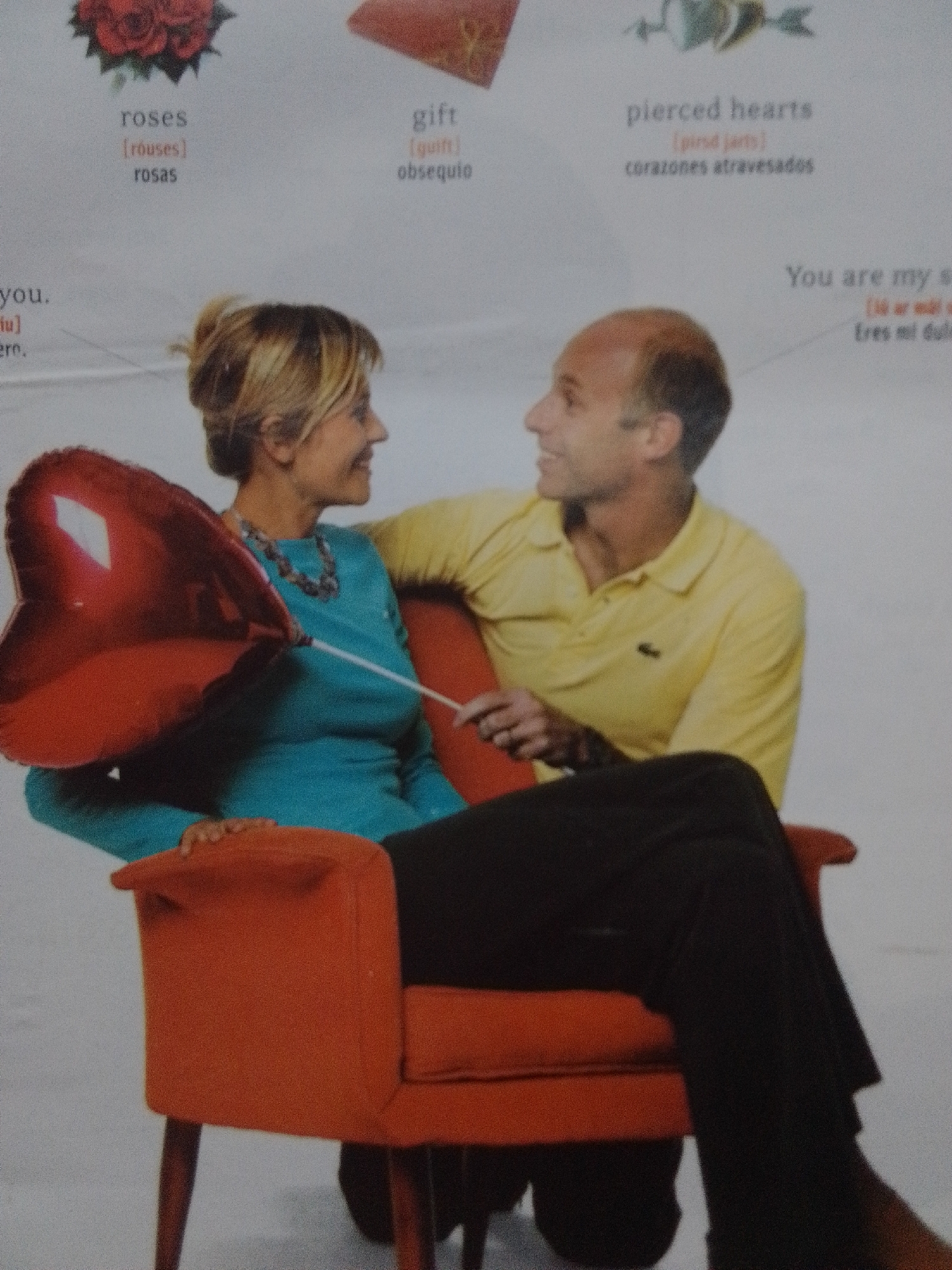This is a medium shot of a man and a woman sitting on an orange armchair. The woman is on the left, holding a large, red, heart-shaped balloon. She is wearing a teal-colored top and a beaded necklace. Her hair is blonde and pulled back. The man is on the right, wearing a yellow polo shirt. He is looking to his left, and the woman is looking forward. Both appear to be smiling. The background is a plain, light-colored surface, likely a wall or studio backdrop.
Visible text in the image includes "roses," "gift," "pierced hearts," and "You are my s." These appear to be labels or captions related to Valentine's Day themes, with some text in Spanish, such as "rosas" (roses) and "corazones atravesados" (pierced hearts). The context provided for La Tagua, Venezuela, is not visually evident in the image itself. The overall mood is romantic and cheerful.
