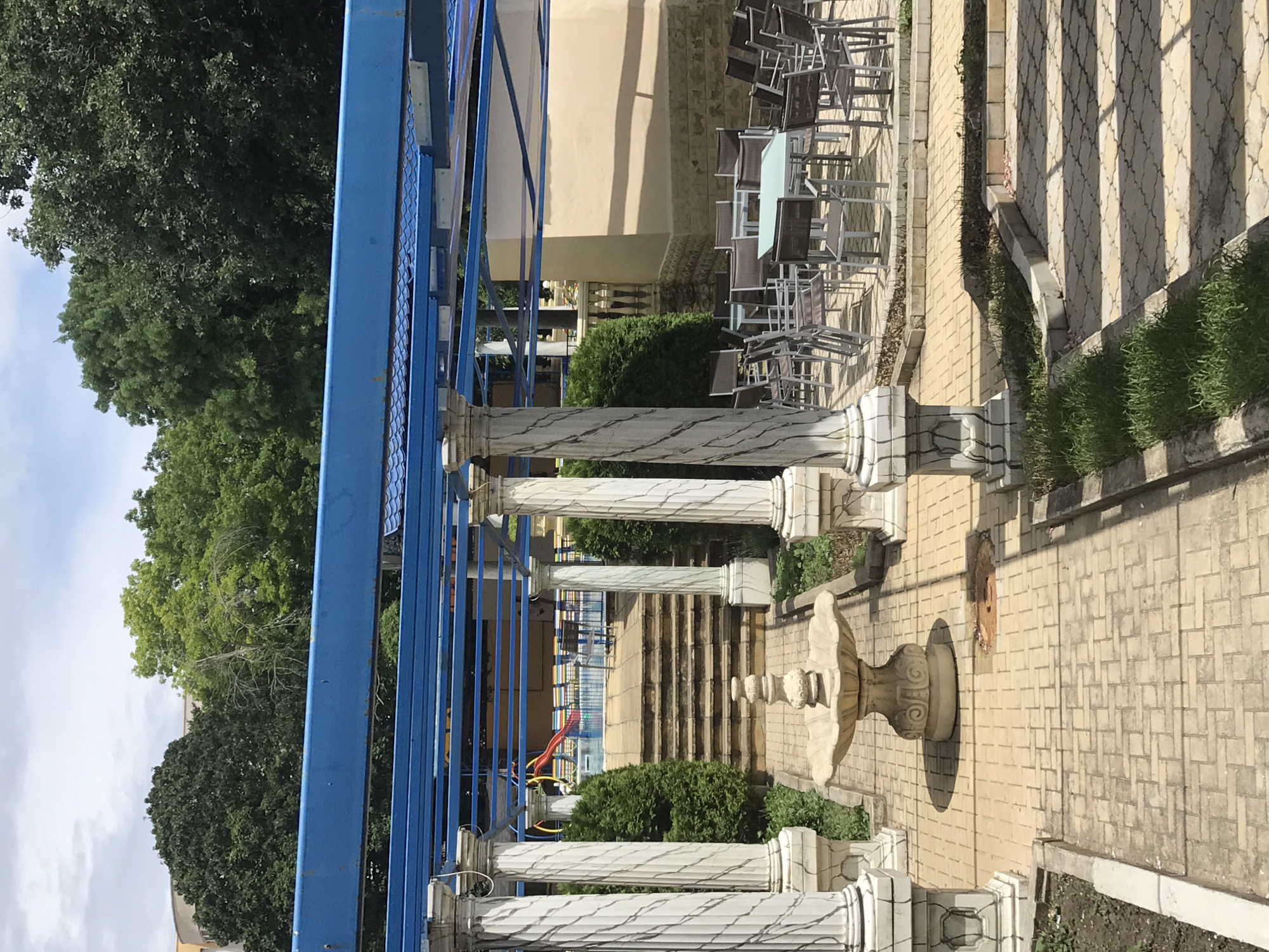 The image depicts an outdoor seating area with a pool in the background, likely located at the Aquapark Aquapolis in Varna, Bulgaria. The foreground features a paved walkway leading to a stone fountain. Flanking the walkway are four large, decorative pillars supporting a blue pergola structure. Beyond the fountain, a set of stone stairs ascends towards the pool area, which is visible with lounge chairs and a red slide. The seating area is furnished with numerous metal tables and chairs. Lush green trees surround the scene, and the sky is partly cloudy.