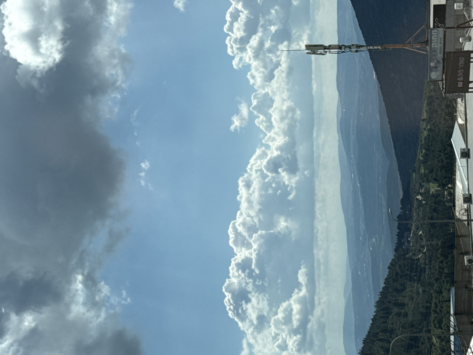 The image presents a wide outdoor vista overlooking a mountainous landscape under a partially cloudy sky. The upper half is dominated by a clear blue sky transitioning to a band of bright, white cumulus clouds stretching across the mid-horizon. Darker, grey clouds occupy the upper left quadrant. Below the clouds, multiple layers of green and blue-grey mountains and hills recede into the distance, indicating a deep valley. The closest hills are densely forested with dark green trees. In the foreground, on the right, a tall cellular transmission tower equipped with multiple antennas is visible. Beneath the tower, partial rooftops of buildings are present, featuring several white HVAC units. A brown sign with Hebrew text is partially discernible, reading "מתוק" (Matok) at the top and "שוקל 6.90" (Shokel 6.90) below. On the lower left, a section of another building and the upper part of a street light are visible. The vantage point is elevated, consistent with a view from a hillside or mountain in the city of Safed, Israel, looking over the surrounding Galilee region. No individuals are depicted.