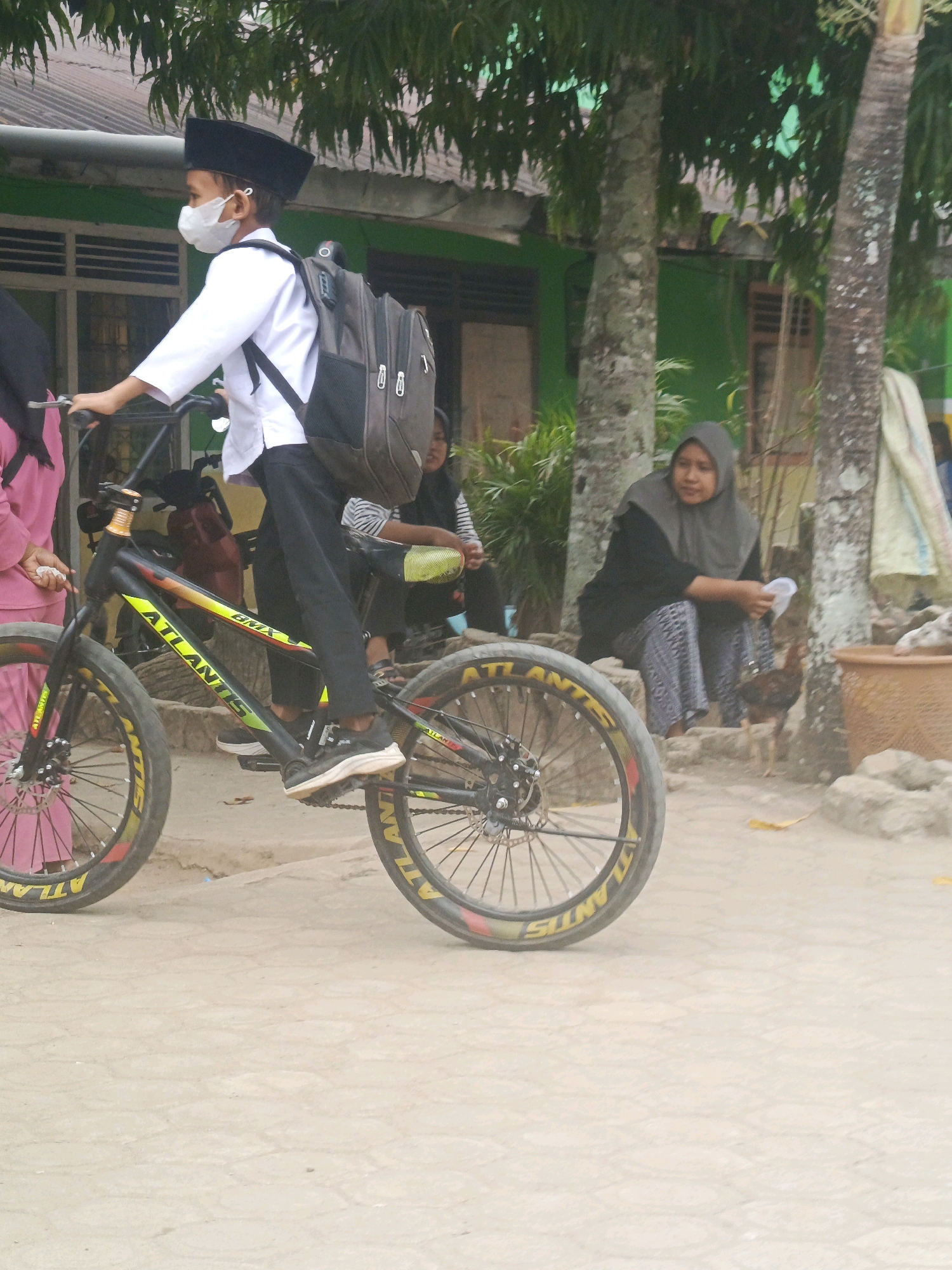 In Megang Sakti II, Indonesia, a young boy wearing a white shirt, black pants, a peci, and a face mask rides a black and yellow BMX bicycle. He is carrying a large, gray backpack. A woman in a gray hijab and patterned pants sits on stone steps to the right, holding a white plastic bag. Another person in a pink outfit is partially visible on the left, next to the boy. The background features trees and a green building, suggesting an outdoor setting near a school or community area. The ground is a light-colored, paved surface. A chicken is visible near the woman on the steps.