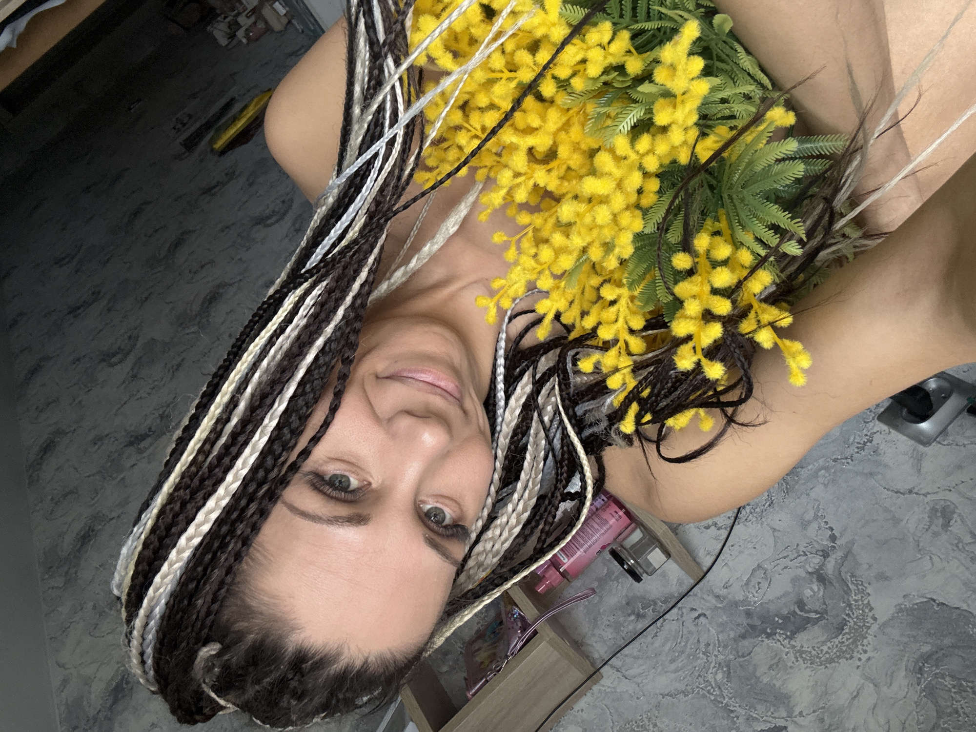 A woman with black and white braided hair is holding yellow flowers. She is indoors. The location and city of Zhukovo, Russia are not verifiable from the image.