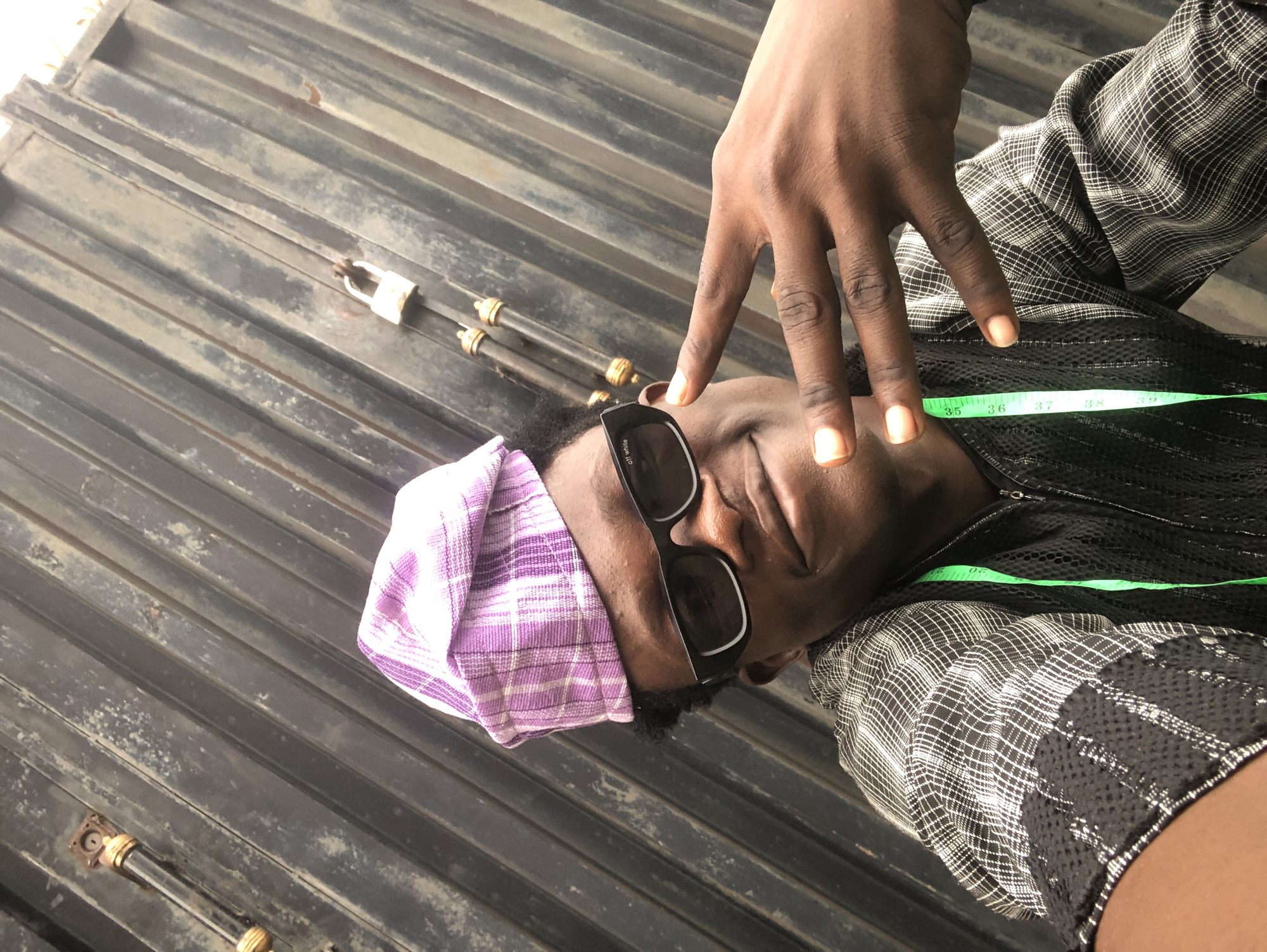 A portrait shows a young Black man, wearing a purple plaid cap, black sunglasses, and a black and white checkered shirt with a green measuring tape visible around his neck. He is positioned in front of a dark, corrugated metal fence, which has a padlock secured to it. The man is smiling and holding up his hand in a peace sign. The overall lighting suggests it is daytime, outdoors. The location is identified as Ado, Nigeria.