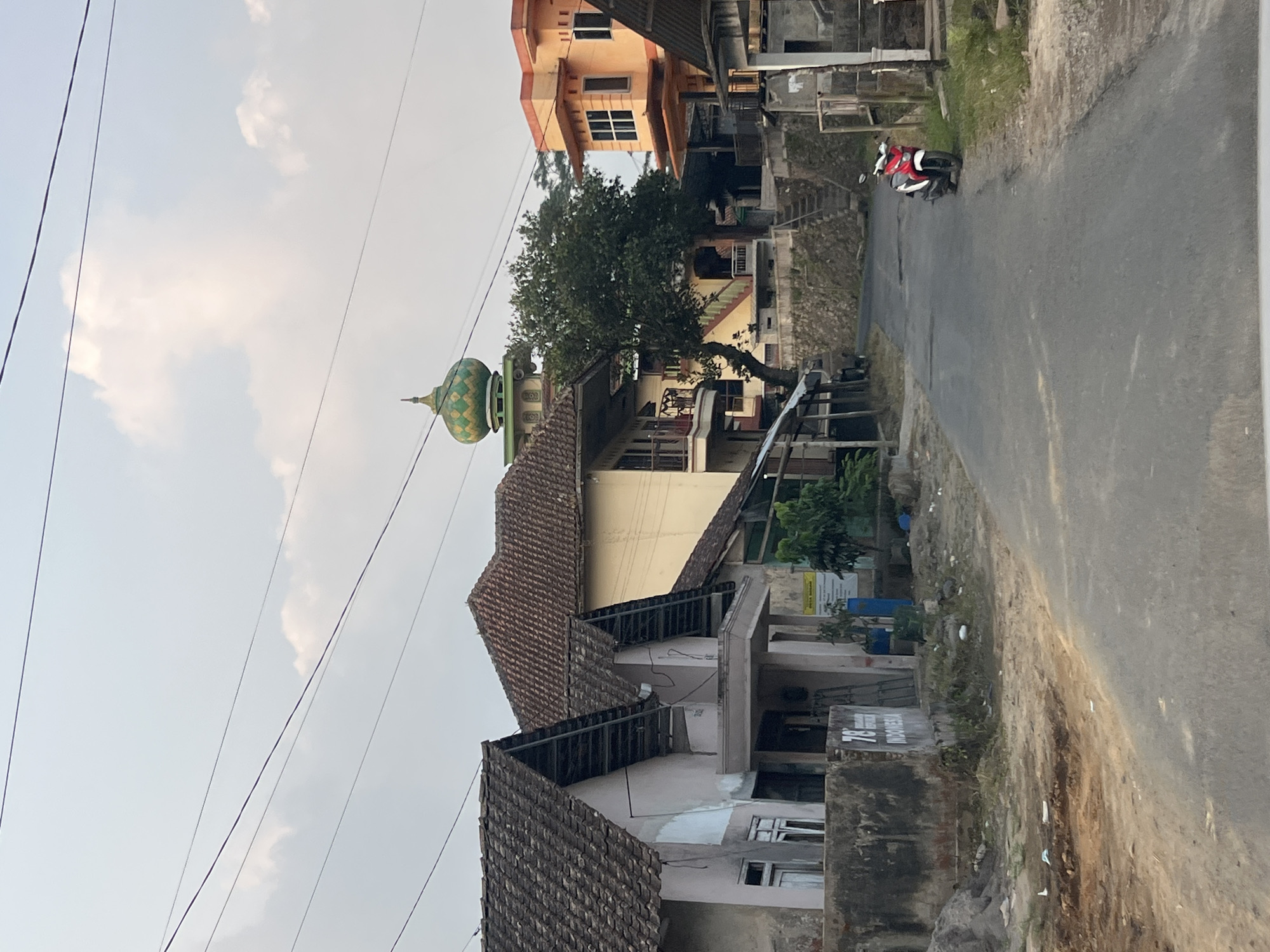 The image depicts a street scene in Beran, Indonesia, characterized by a mix of residential buildings and a prominent mosque. The foreground shows an asphalt road with a red and white motorcycle parked on the right side. To the left of the road, there are two aged, light-colored residential structures with tiled roofs. One building features a concrete wall with the number "78" and the word "INDONESIA" stenciled on it. Behind these houses, a larger, light yellow building with balconies is visible. Further in the background, the distinctive green and gold dome of a mosque rises above the trees and other buildings. The sky is overcast with scattered clouds, and overhead power lines crisscross the frame. The overall impression is of a rural or semi-urban Indonesian village environment.