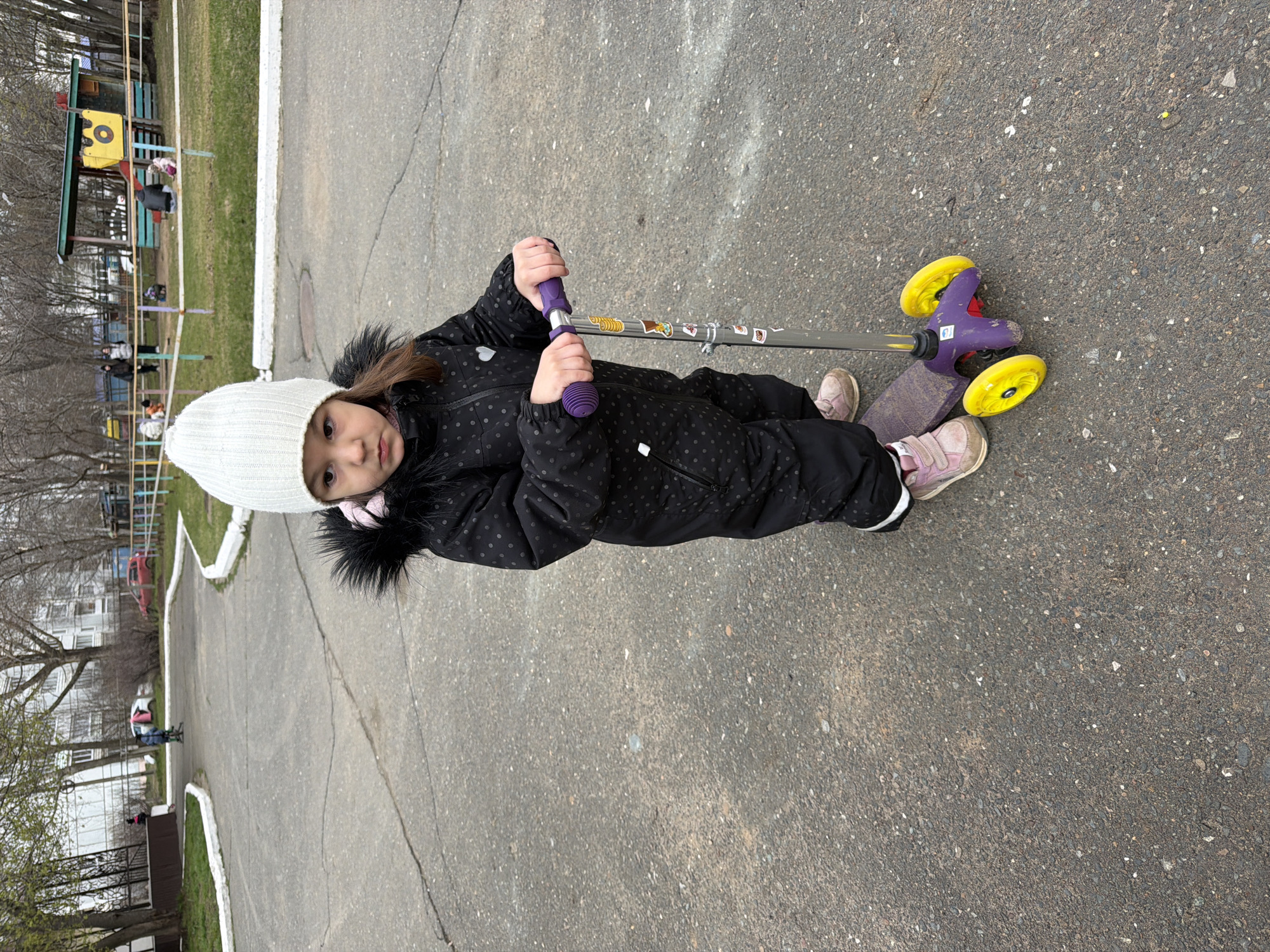 A young child in a black winter jumpsuit and white knit hat stands on a gray asphalt surface with cracks. The child is holding a purple and yellow scooter. The child is looking towards the camera. In the background are trees, grass, and a playground structure. A white building is also visible in the background. The location is outdoors in the city of Chaikovsky, Russia.