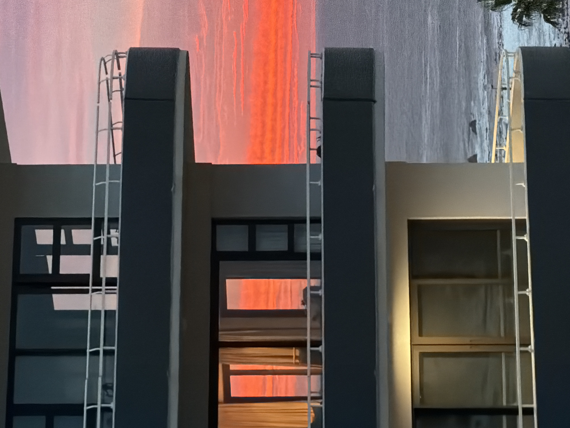 The image depicts a building facade in Cape Town, South Africa. The architecture features two dark gray rectangular pillars with white ladder-like structures attached to the exterior. Between the pillars, windows are visible, reflecting an orange and pink sky suggestive of a sunset. To the left, a window shows a darker, blue-gray sky. The building's surfaces are a light gray or beige color. The windows are divided into rectangular panes. The overall composition of the image is symmetrical, focusing on the contrast between the architectural elements and the sky.