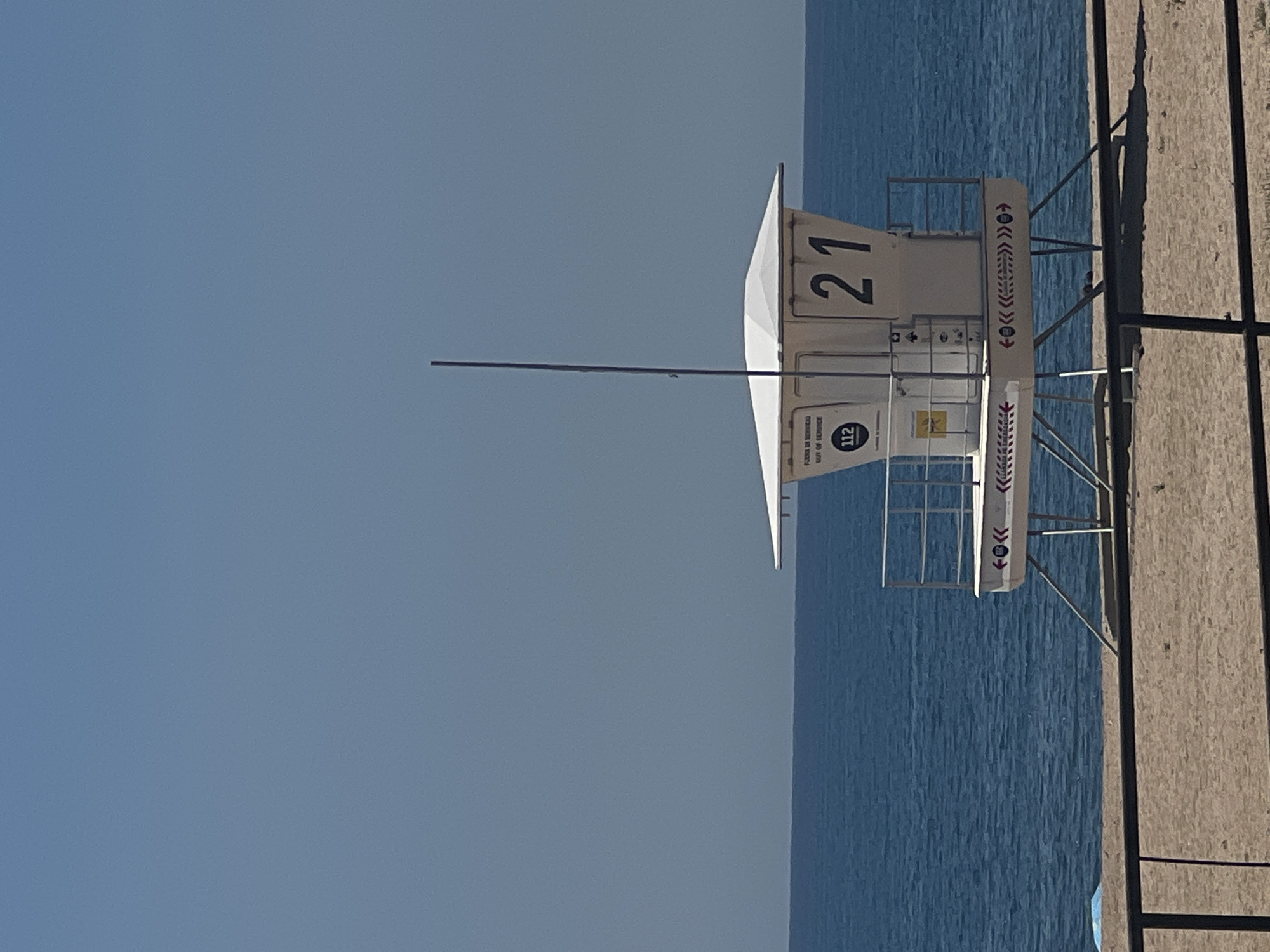 The image captures a lifeguard tower on a beach, most likely in Sant Pol De Mar, Spain. The tower, marked with the number "21", has a white body with the inscription "112" and decorative arrow motifs. It stands on stilts, with a ladder providing access. The sky is a clear, pale blue. The sea is visible beyond the tower, appearing calm. A portion of the sandy beach is visible to the right. A long, thin metal pole extends horizontally from the tower. The overall scene is bathed in bright daylight.