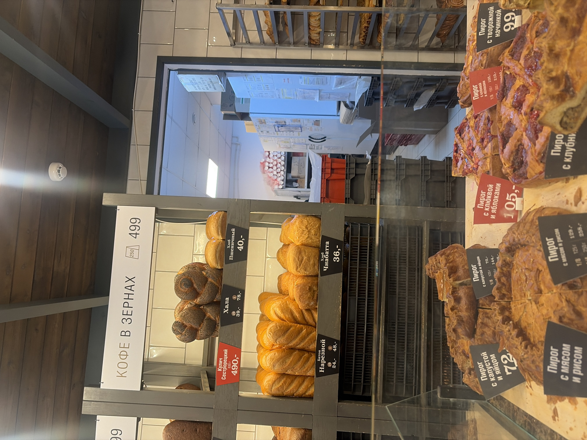This image shows a bakery display case in Saint Petersburg, Russia. Shelves are filled with various bread and pastries. Signs indicate prices in rubles for items such as challah bread, wheat bread, ciabatta, sliced bread, and pies with different fillings. In the background, a glimpse of a kitchen area can be seen.